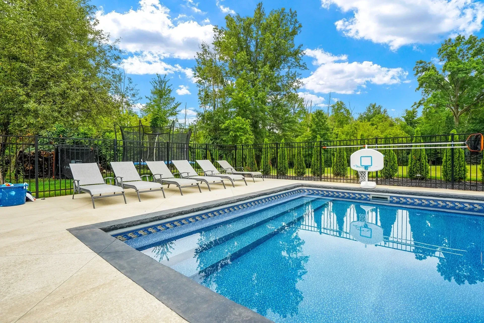 Swimming pool with sun loungers, basketball hoop, and trees under a blue sky.