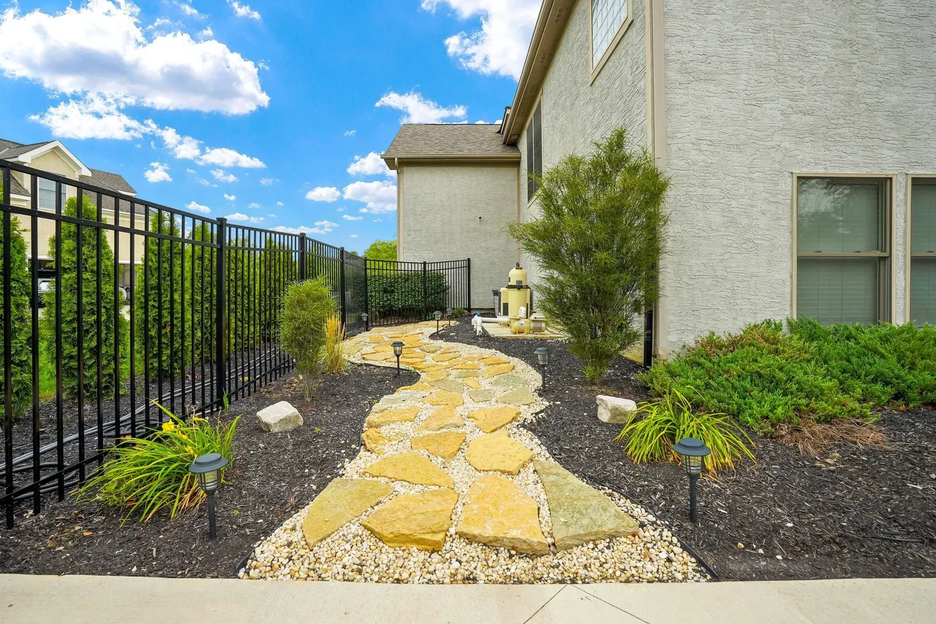 A stone path winds through a landscaped yard next to a light-colored house and a black fence under a blue sky.