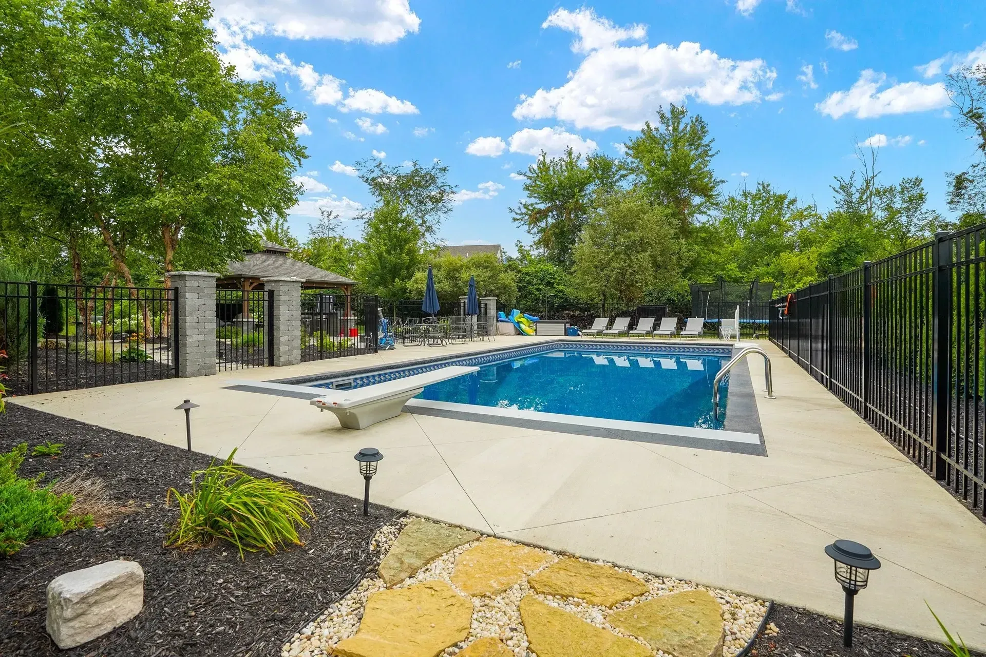 A backyard with a rectangular pool, stone patio, black fencing, and a gazebo under a bright blue sky.