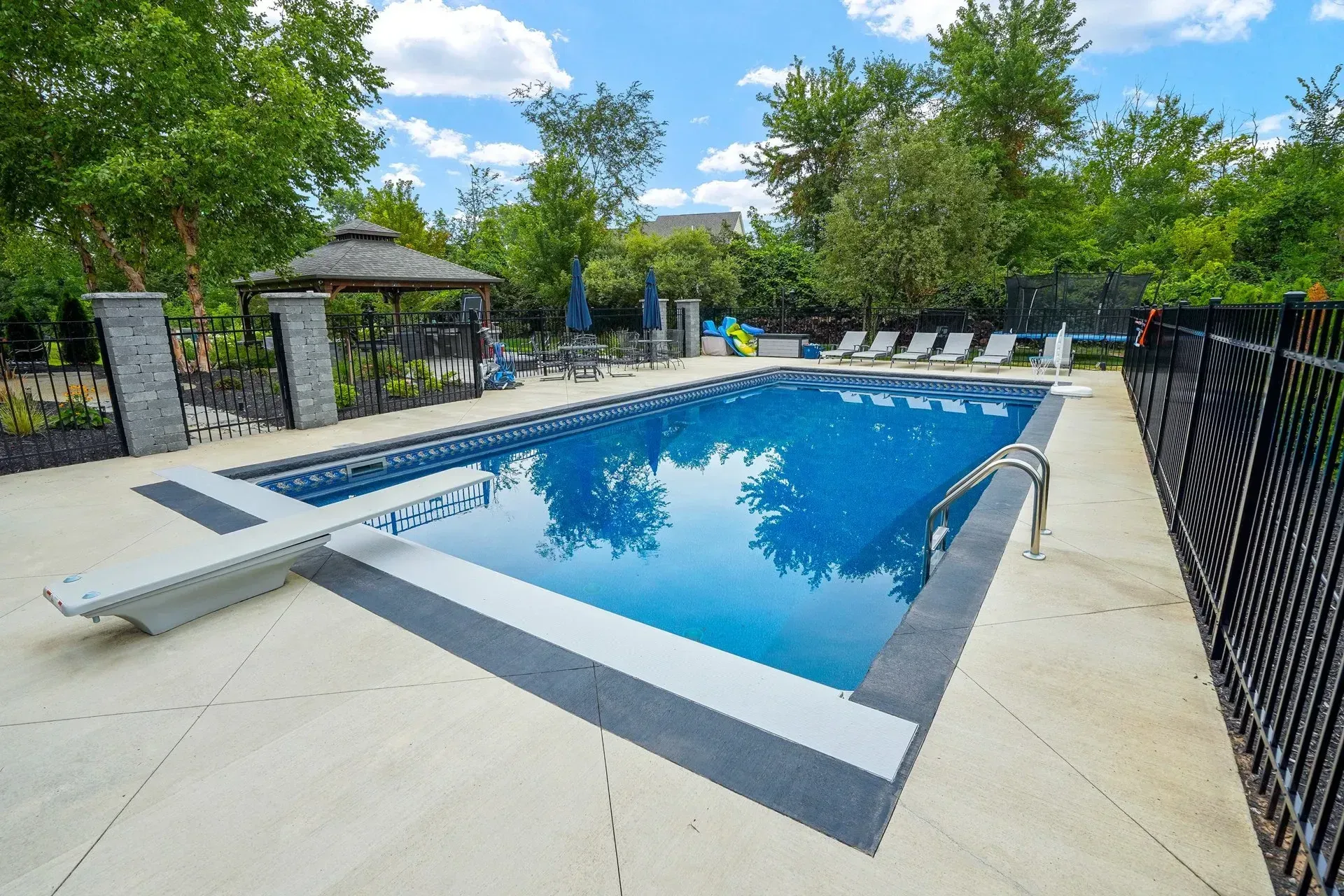 A rectangular swimming pool surrounded by a patio with a diving board, loungers, and a gazebo in a backyard.