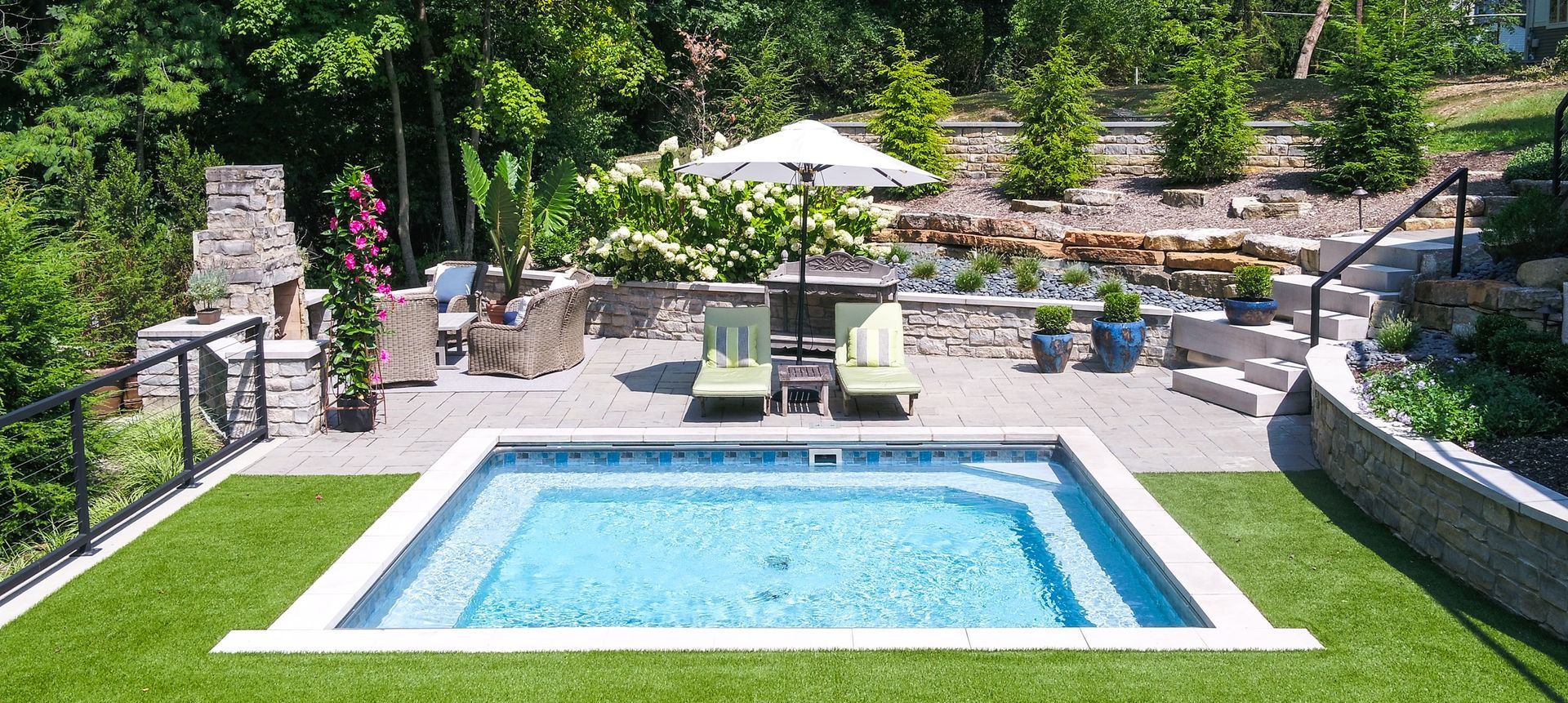 A backyard patio with a small rectangular pool, lounge chairs, and an umbrella. The patio is surrounded by greenery.