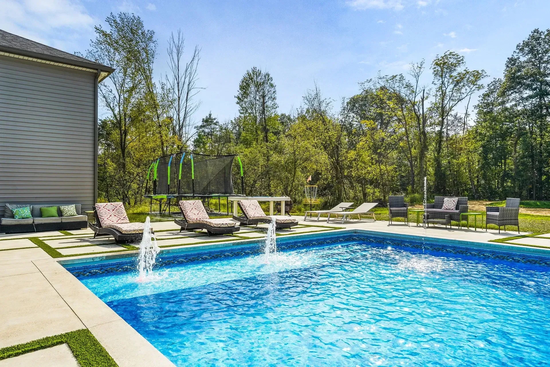 A luxurious backyard pool with fountains, lounge chairs, and a seating area next to a house. Green trees are in the background.