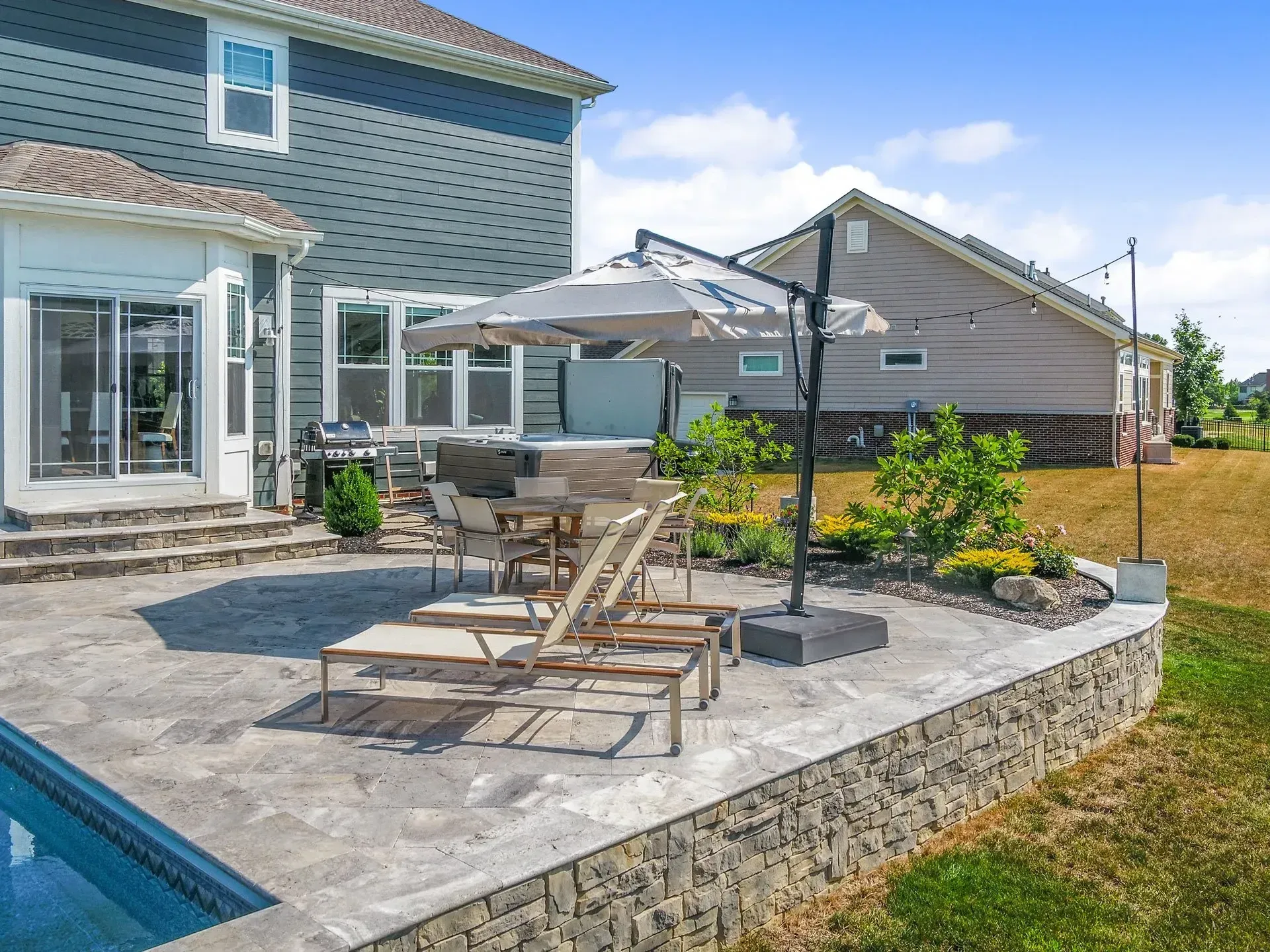Backyard patio with a pool, lounge chairs, and an outdoor kitchen. A raised stone wall borders the patio, with a house and yard in the background.