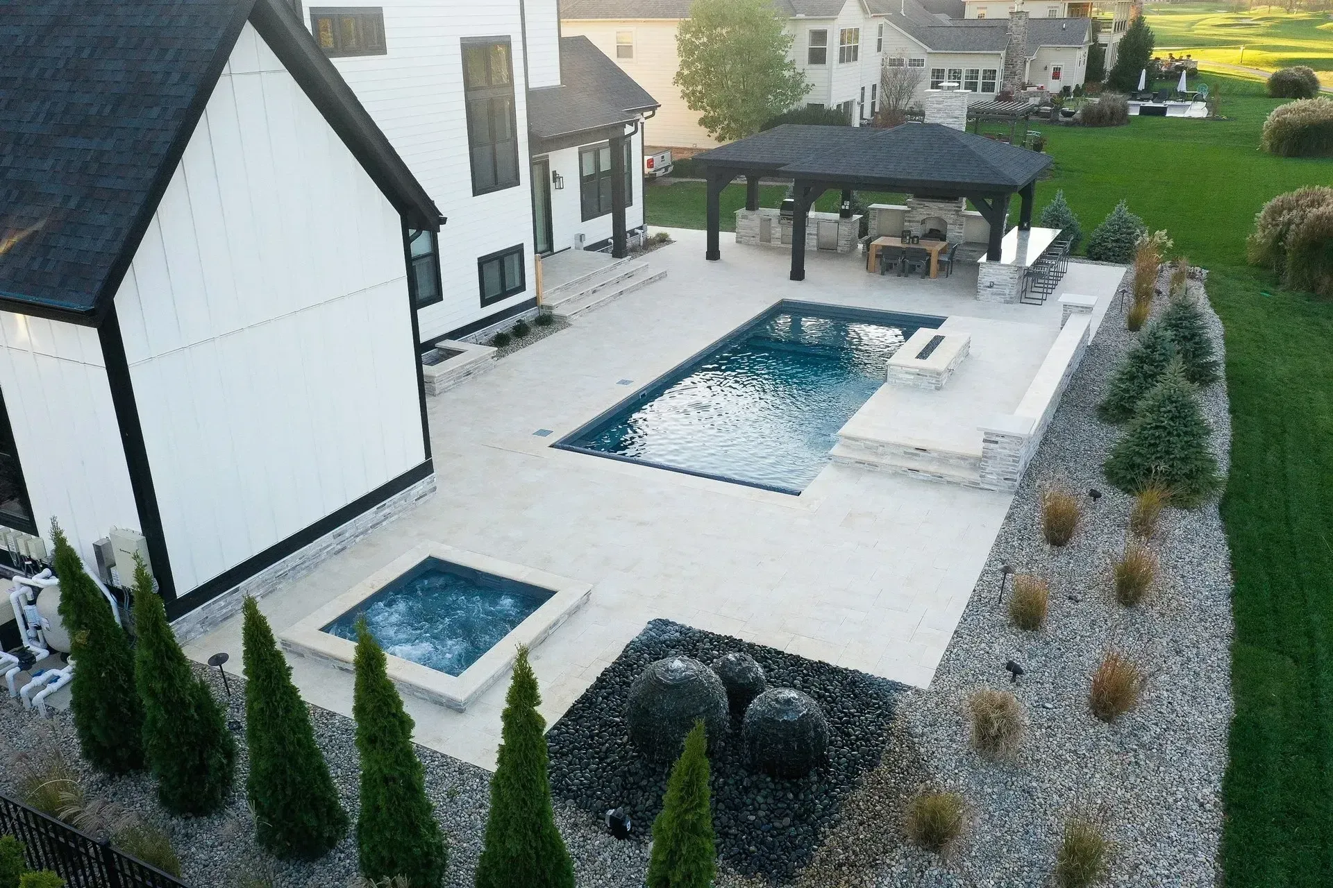 An aerial view of a modern backyard with a pool, spa, patio, and outdoor dining area. The home is white and black with green grass and landscaping.