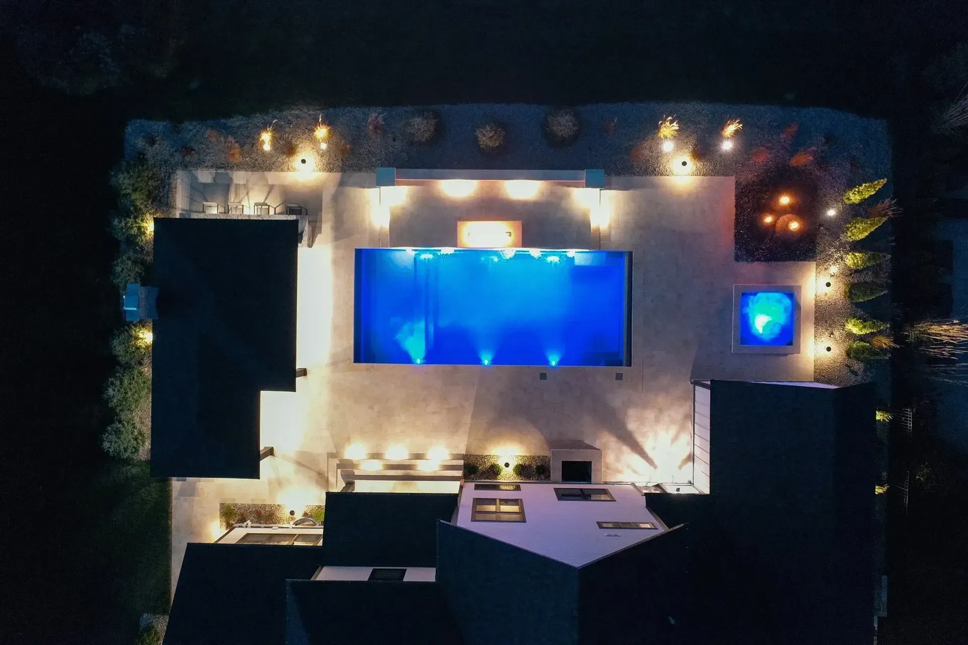 Aerial view of a modern backyard at night with a blue-lit rectangular pool, surrounded by patio, lights, and landscaping.