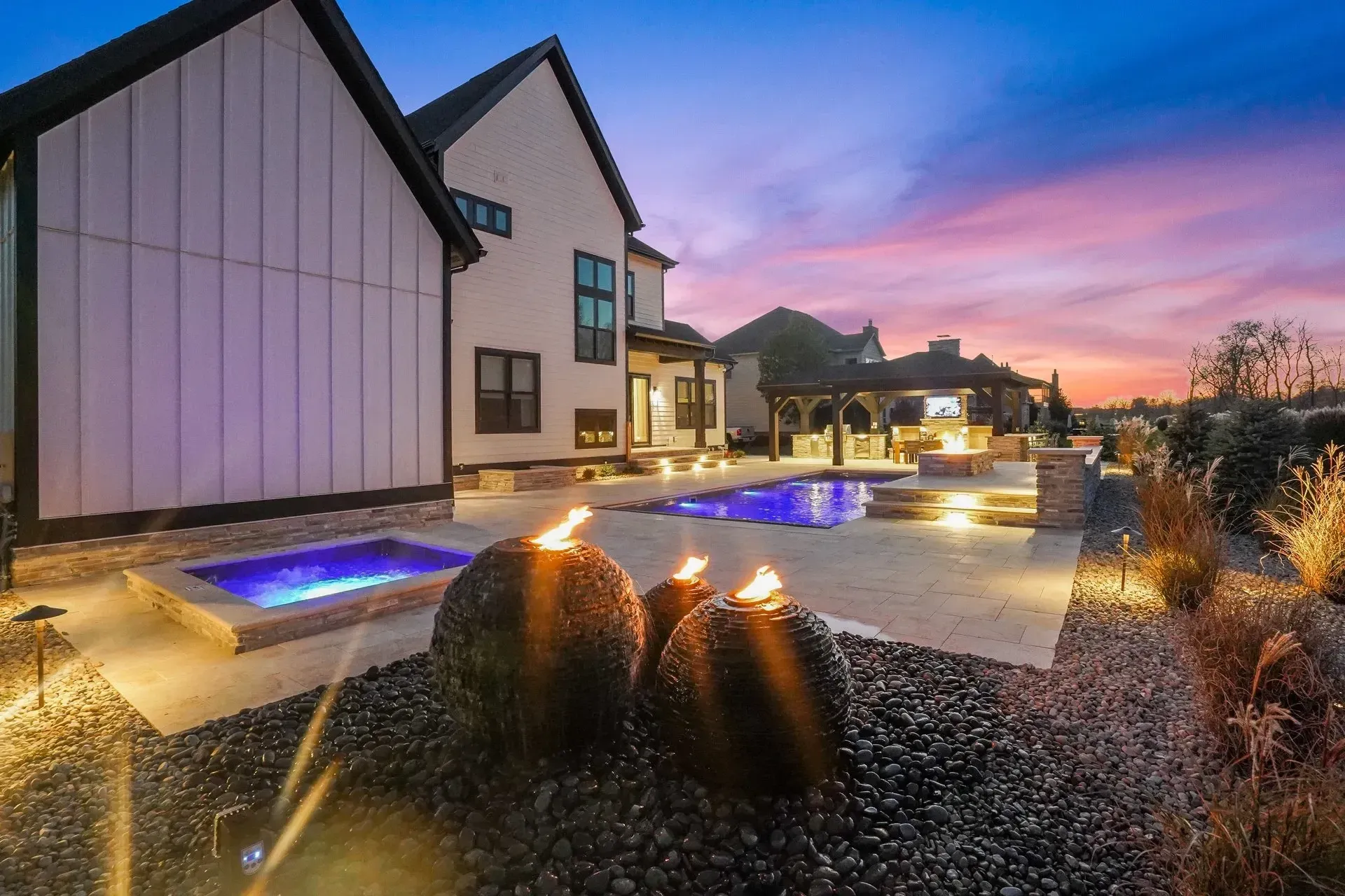 Luxury backyard with a pool, jacuzzi, and fire features, illuminated at dusk with a colorful sky.