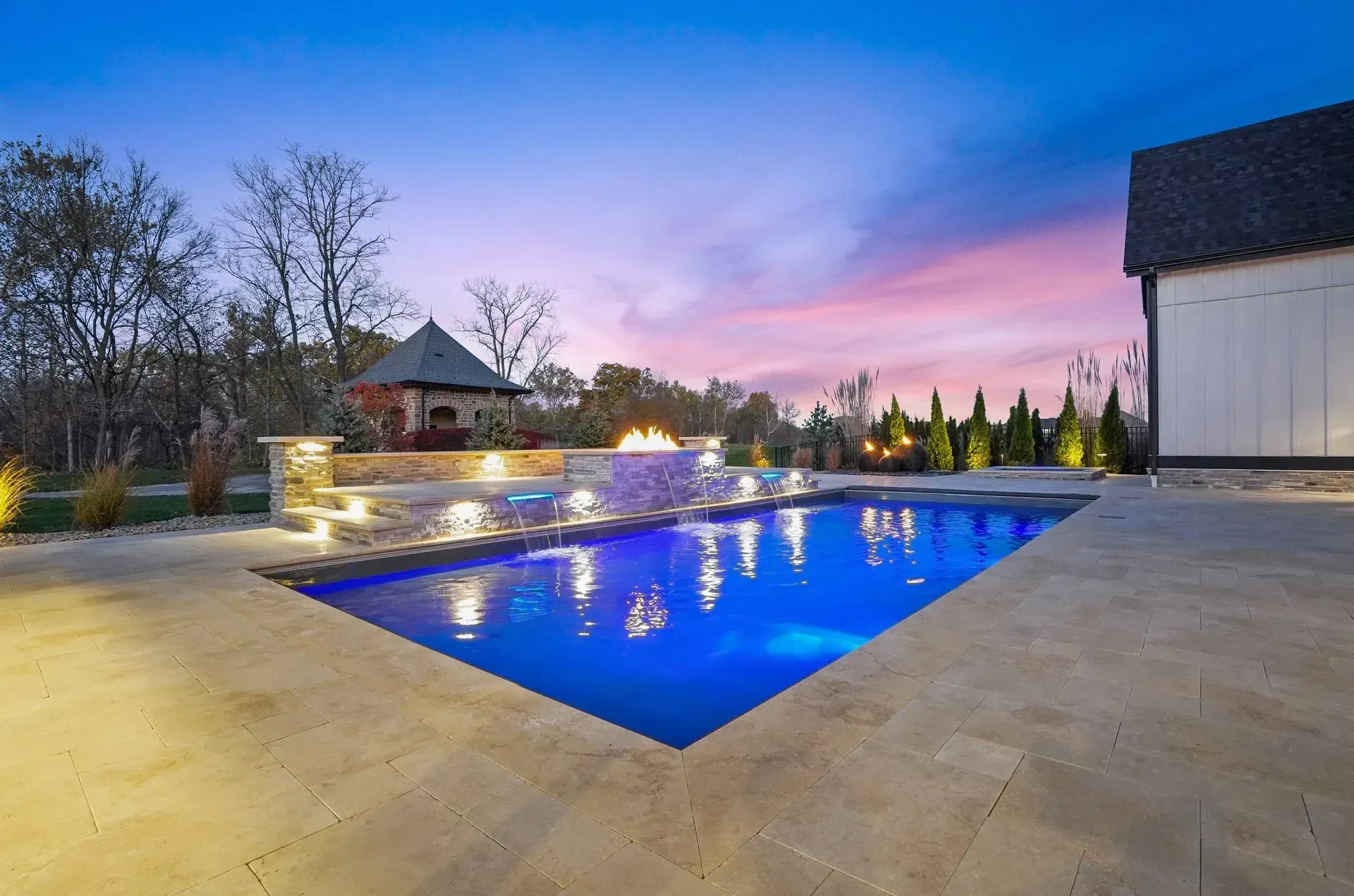 Pool with blue water and a fire pit at dusk. A house is in the background with a colorful sky.