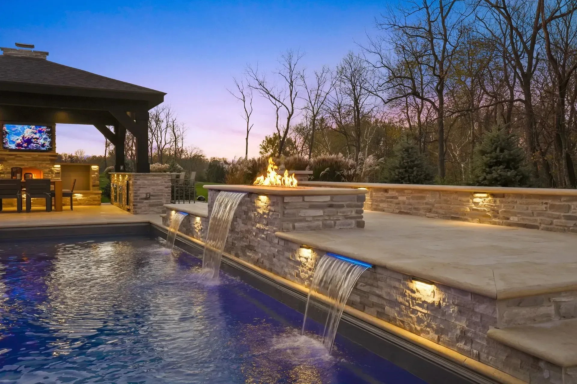 A luxurious outdoor pool with cascading waterfalls, a gazebo with a TV, and a fire pit, all lit up at dusk.