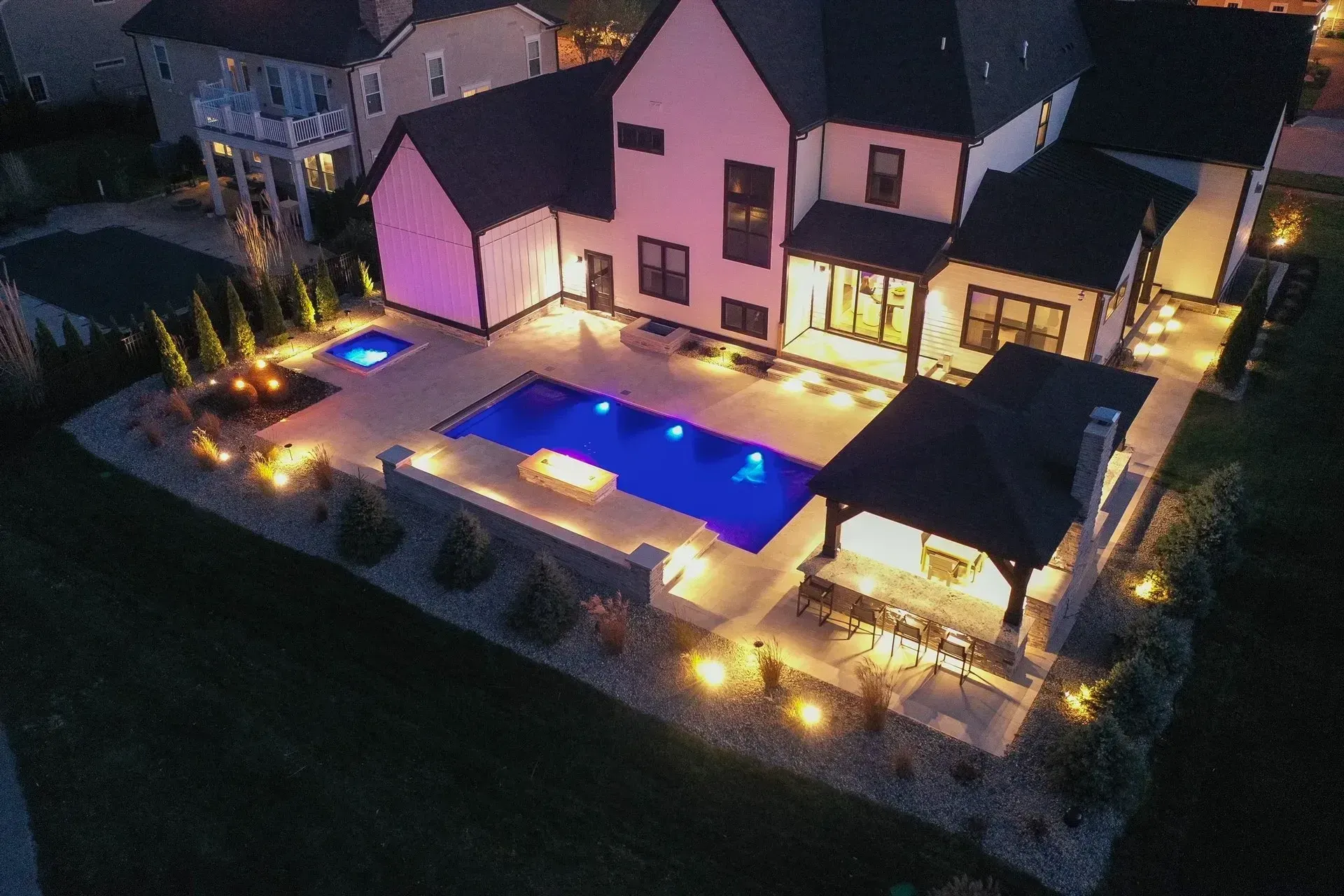 Aerial view of a modern home's illuminated backyard with a pool, hot tub, fire pit, and covered patio. The house is light-colored with dark accents.