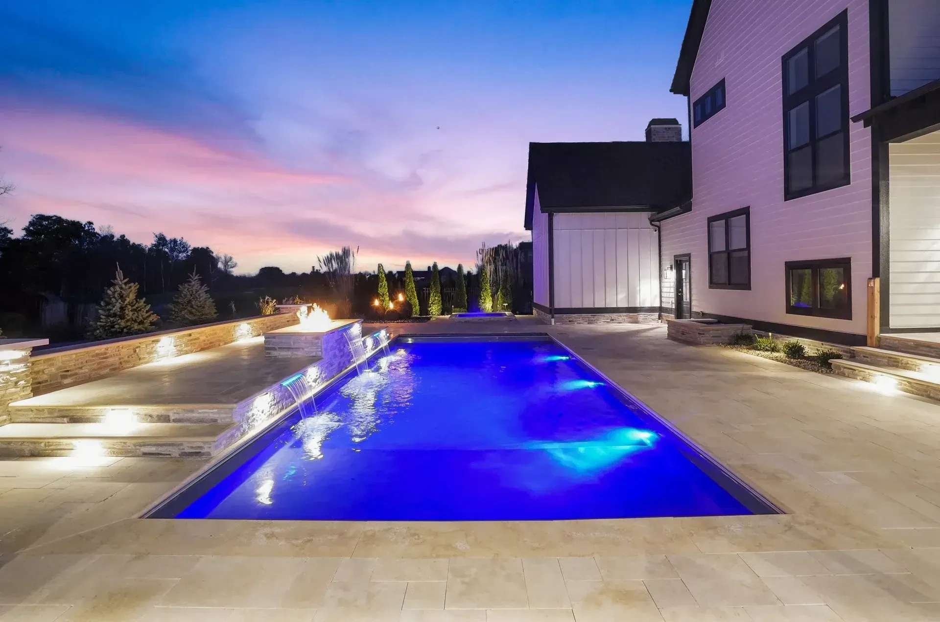 An illuminated rectangular swimming pool with a blue glow sits beside a modern white house at dusk. Stone steps and a fire pit are also visible.