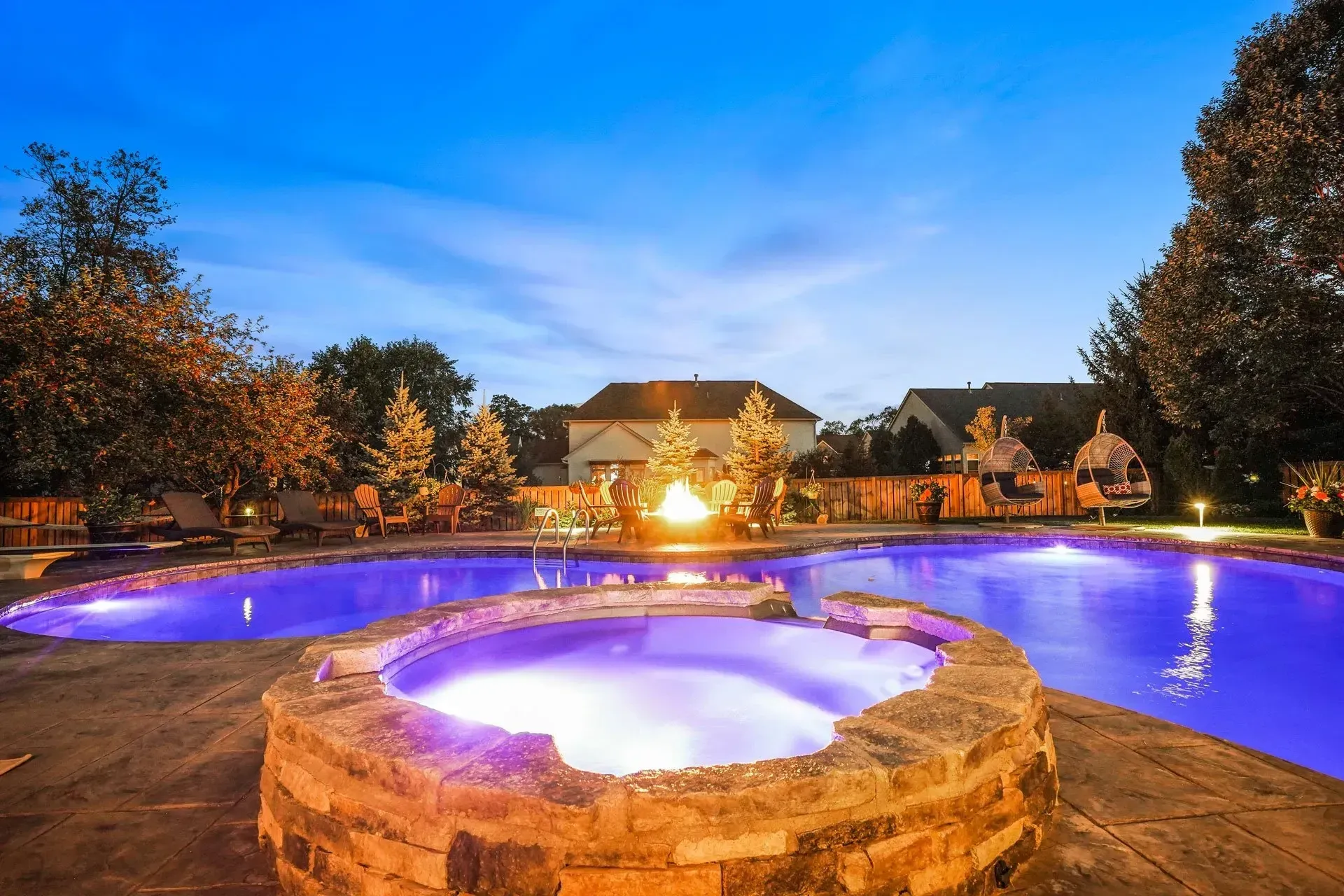 Nighttime backyard with a pool and hot tub illuminated in blue and purple, fire pit burning, and trees.