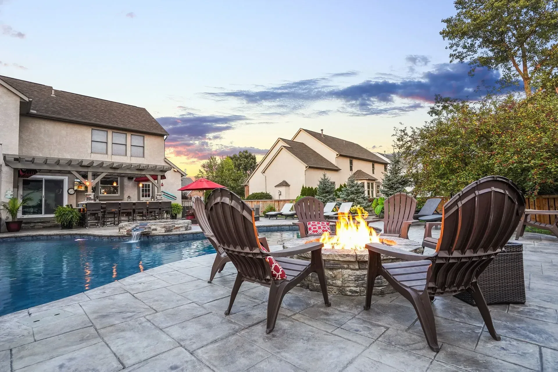 Backyard with a pool, fire pit, and chairs. Houses are in the background. Dusk sky with clouds.