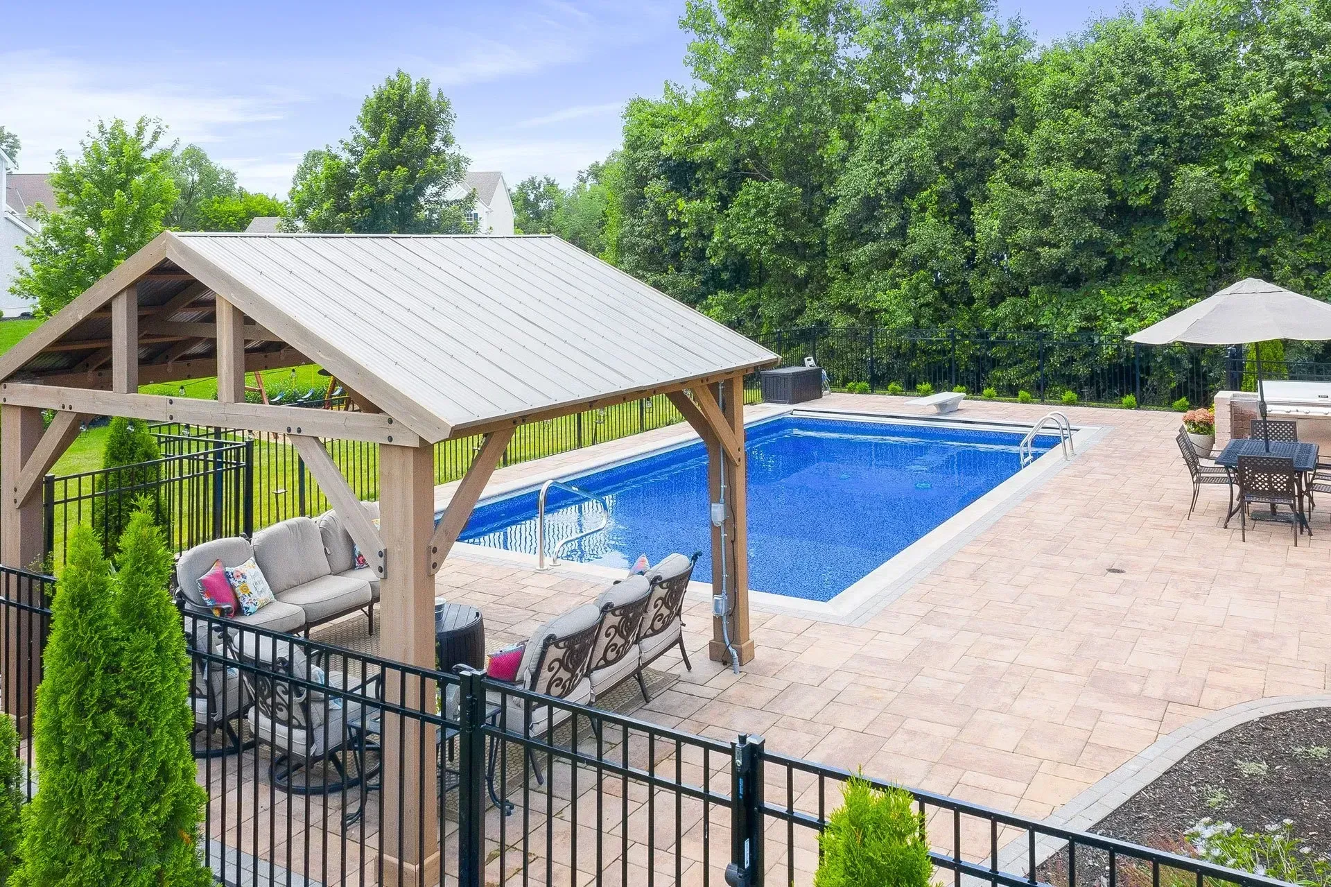 Backyard pool with gazebo, patio furniture, and a black metal fence. Lush green trees and a blue sky in the background.