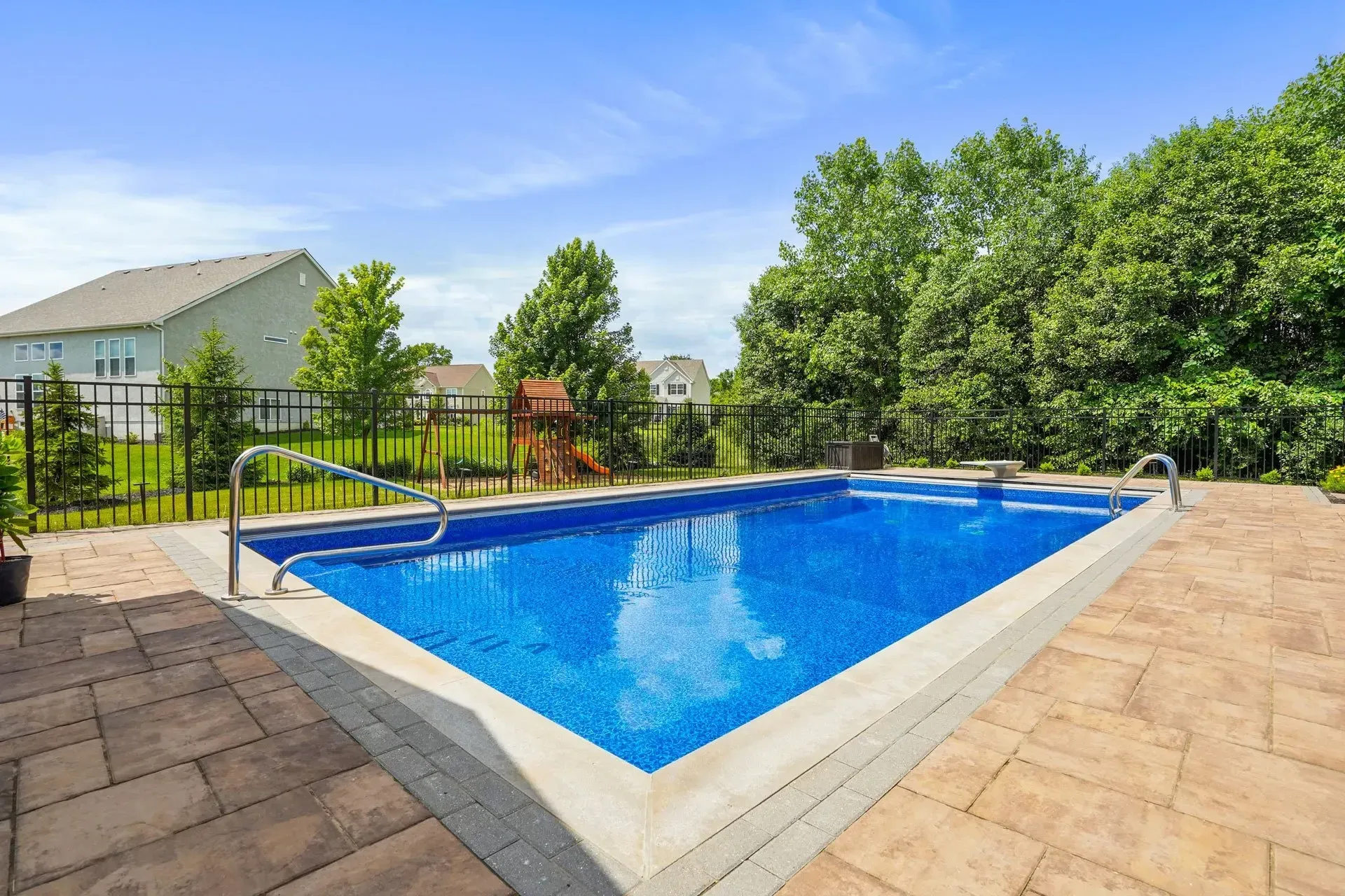 Rectangular in-ground swimming pool with blue water surrounded by brick patio and trees under a blue sky.
