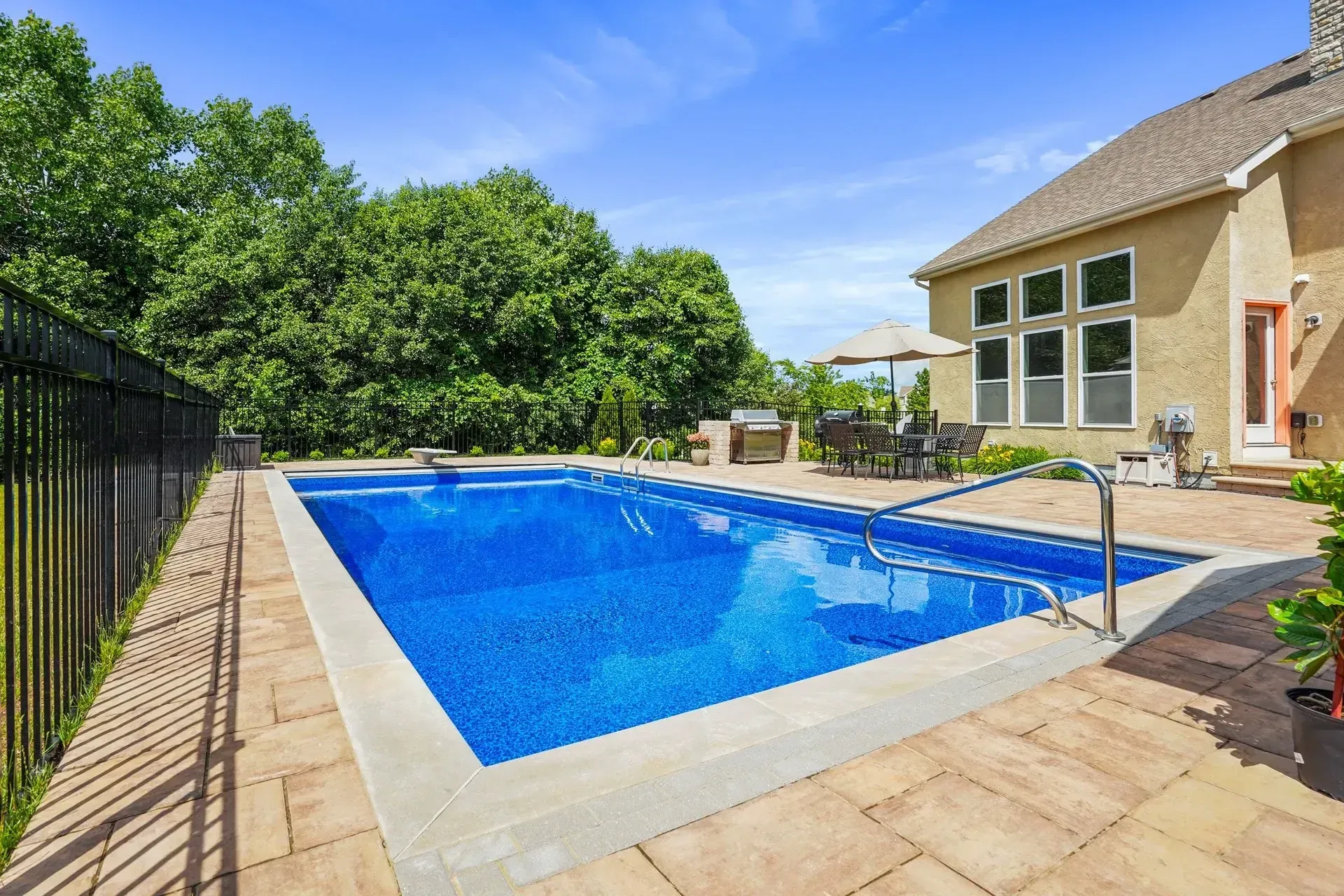 Backyard with rectangular blue pool, patio, dining set, grill, and a house with tall windows under a bright blue sky.