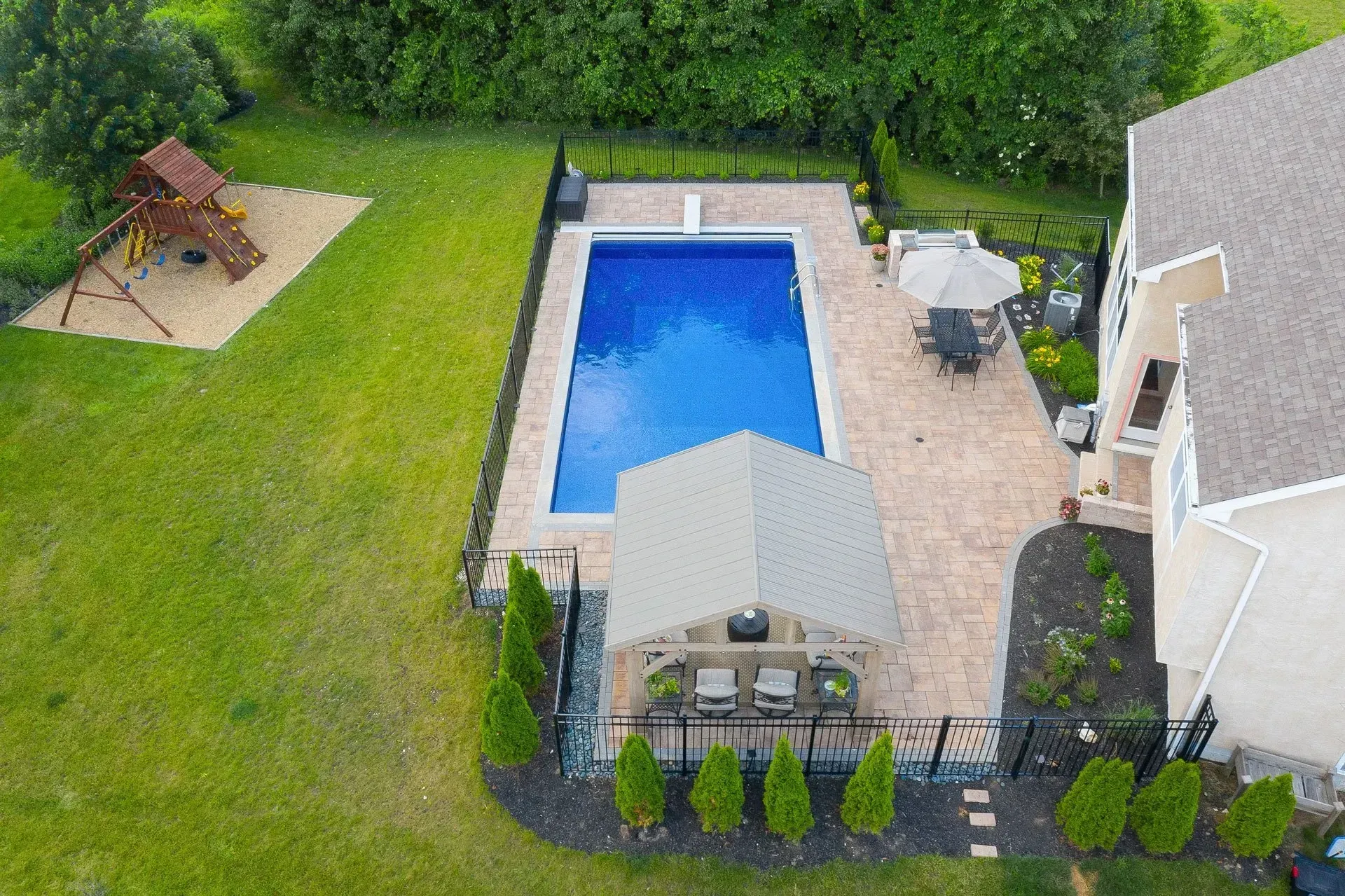 Aerial view of a backyard with a rectangular pool, patio, gazebo, and swing set, surrounded by grass and landscaping.