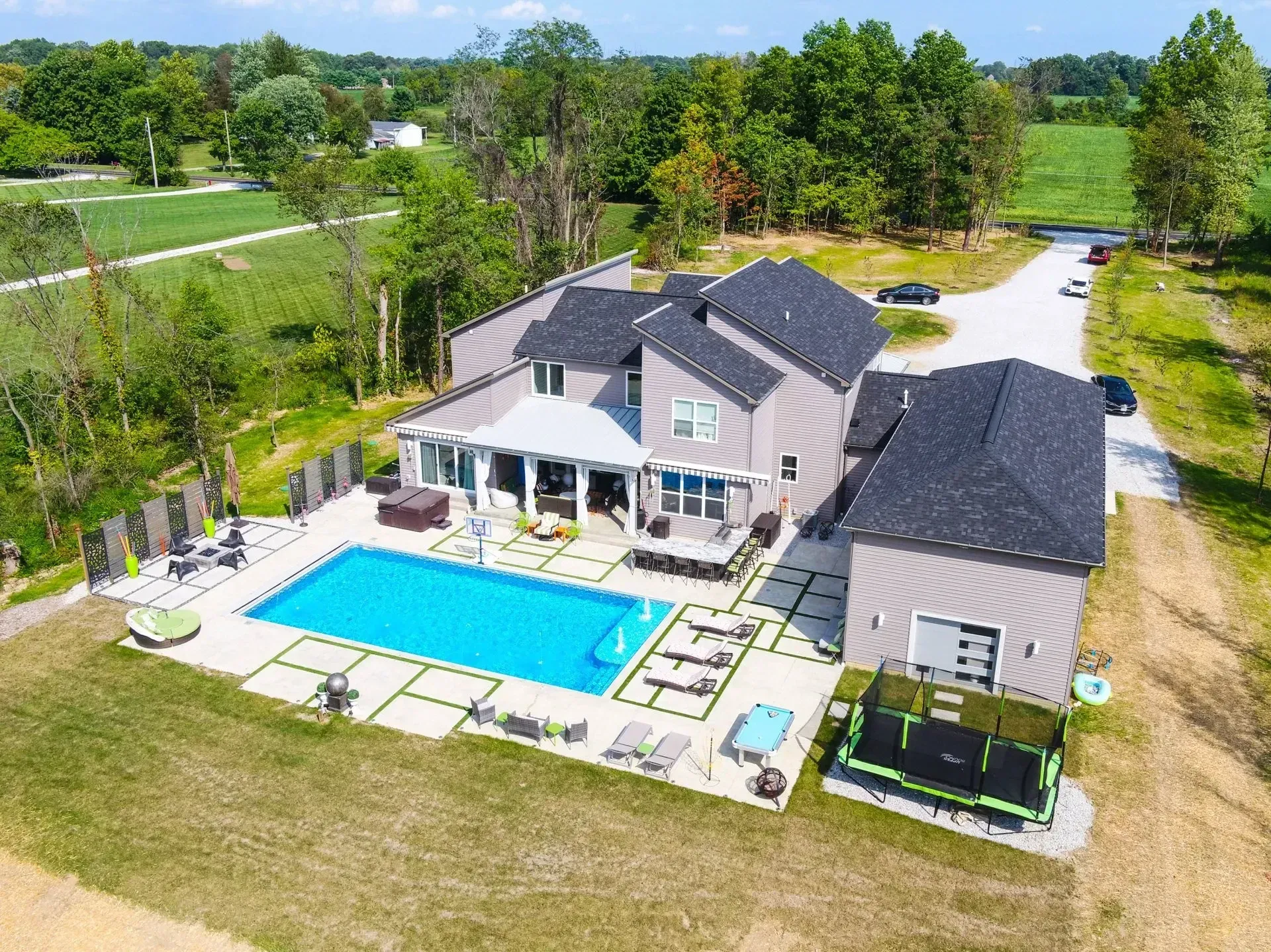 Aerial view of a modern house with a pool, patio furniture, and a trampoline on a large green lawn.