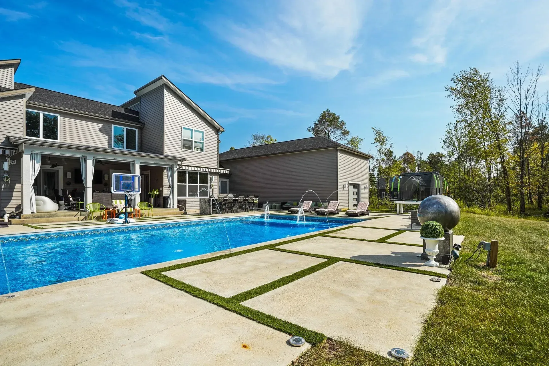 Backyard with pool, patio, and house under blue sky. Includes basketball hoop and landscaping with trees.