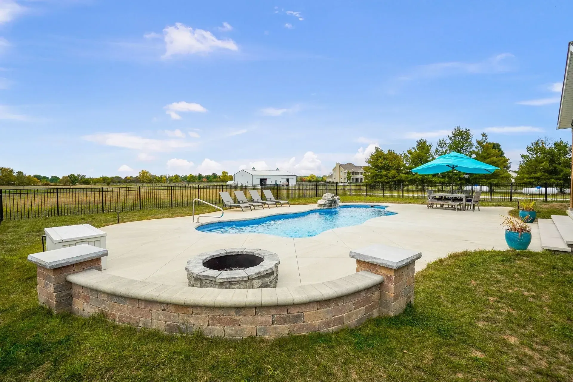 Backyard with a swimming pool, fire pit, and outdoor seating. Sunny day with a blue umbrella and grassy area.