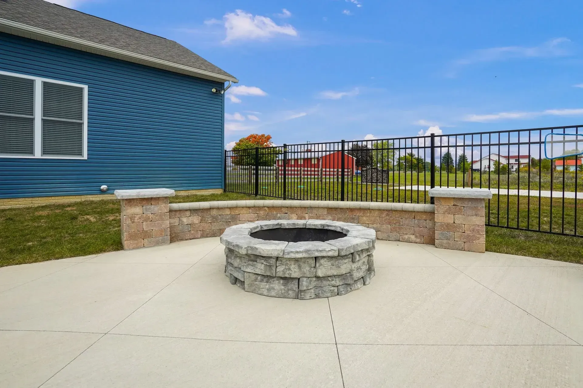 A backyard patio with a stone fire pit, surrounded by a low wall and a black fence. The house is blue, and the sky is clear.
