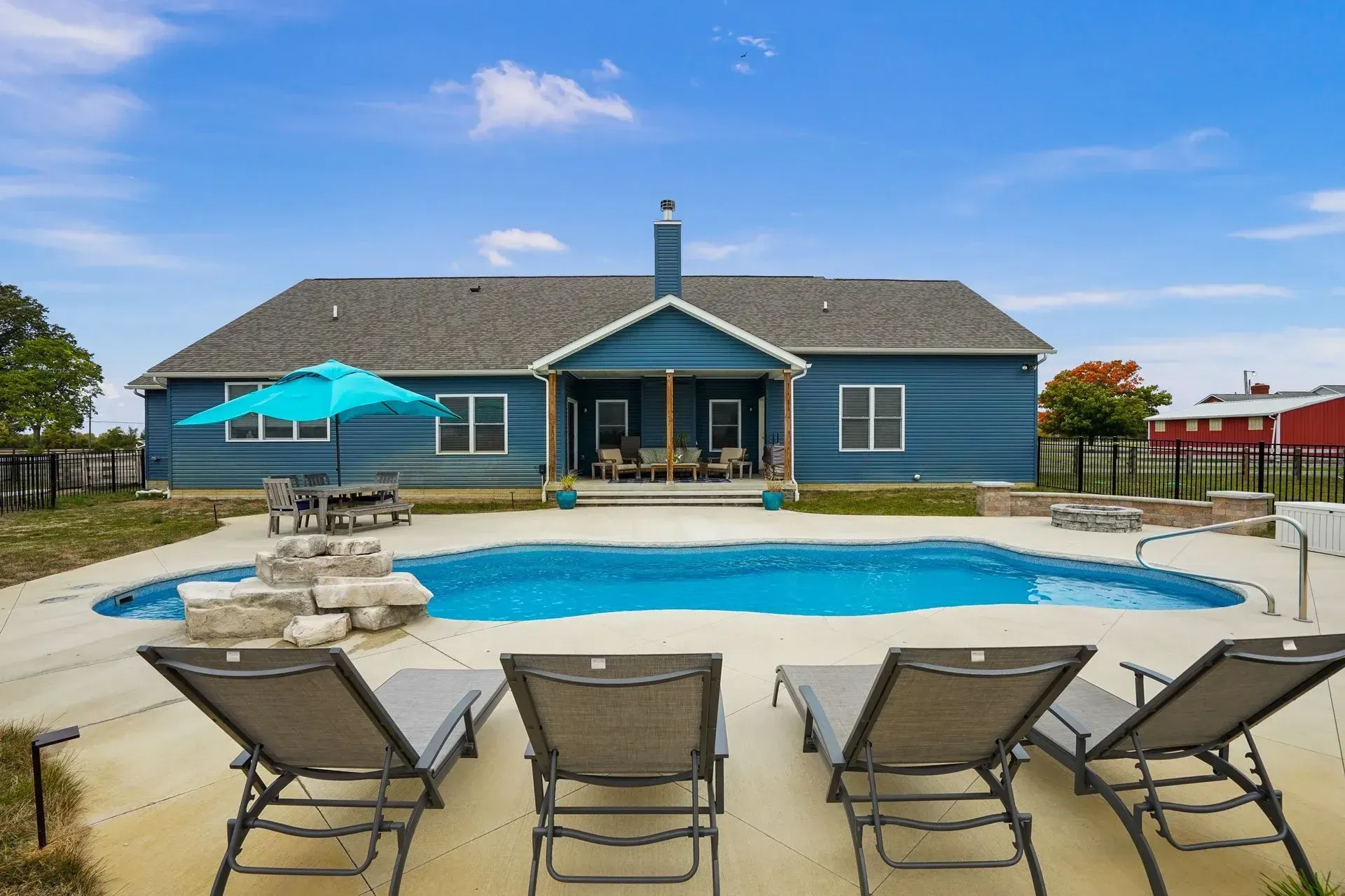 A blue house with a pool in the backyard. There are lounge chairs, a table with an umbrella, and a stone feature.