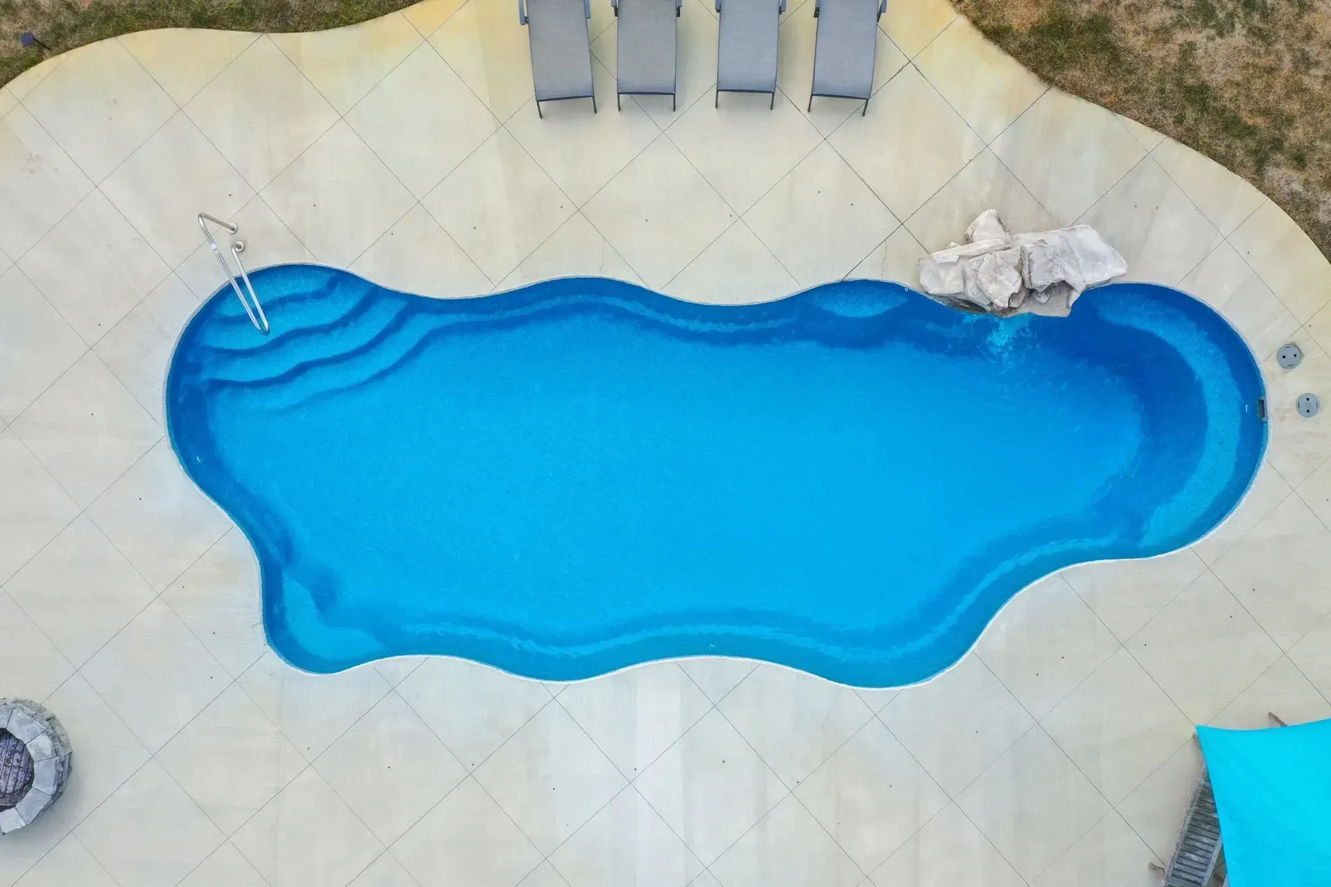 Aerial view of an irregularly-shaped swimming pool filled with blue water. Four gray lounge chairs are visible along one side.