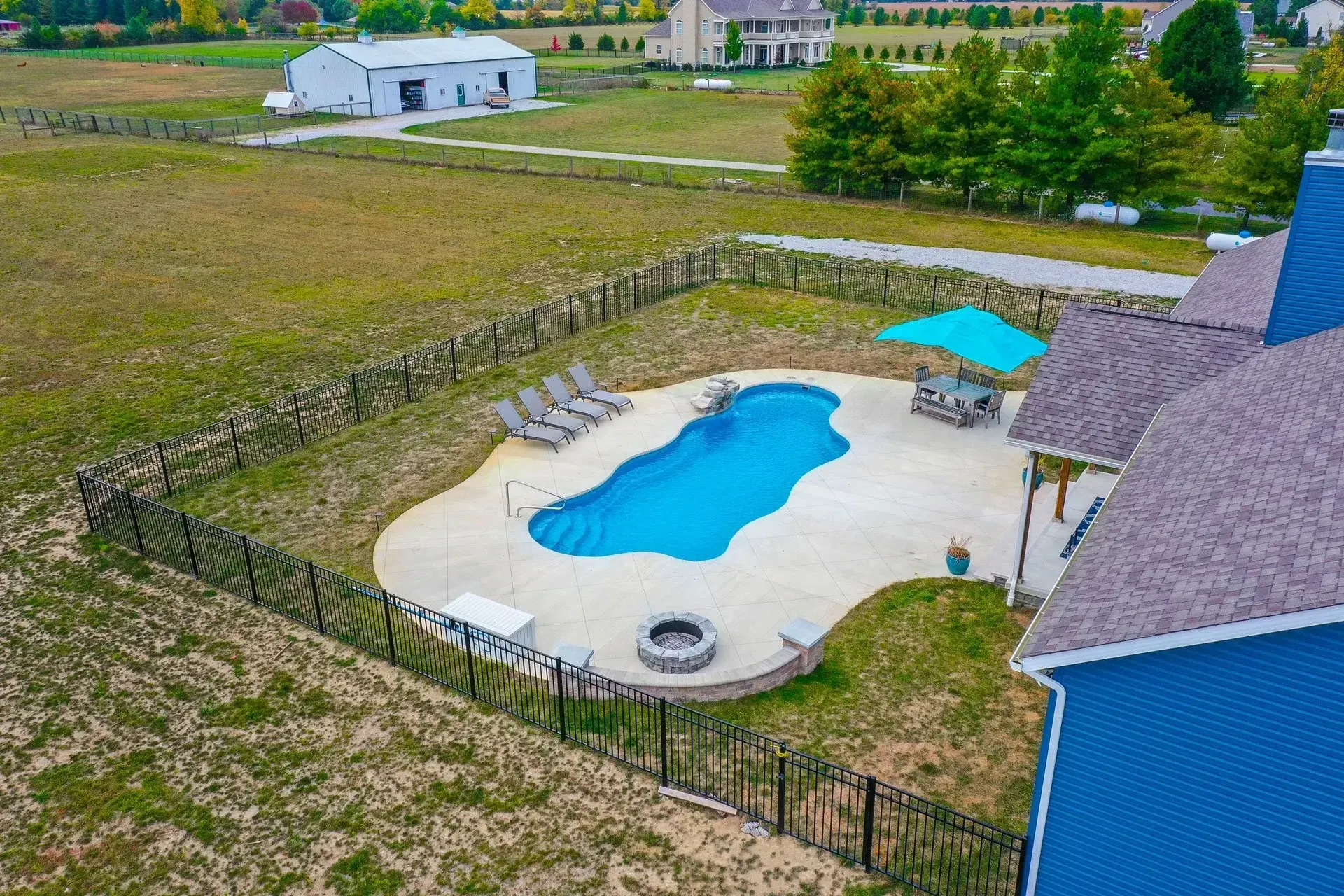 Aerial view of a backyard with a blue pool, patio, and a blue house. The setting is a rural area with a barn and open field.