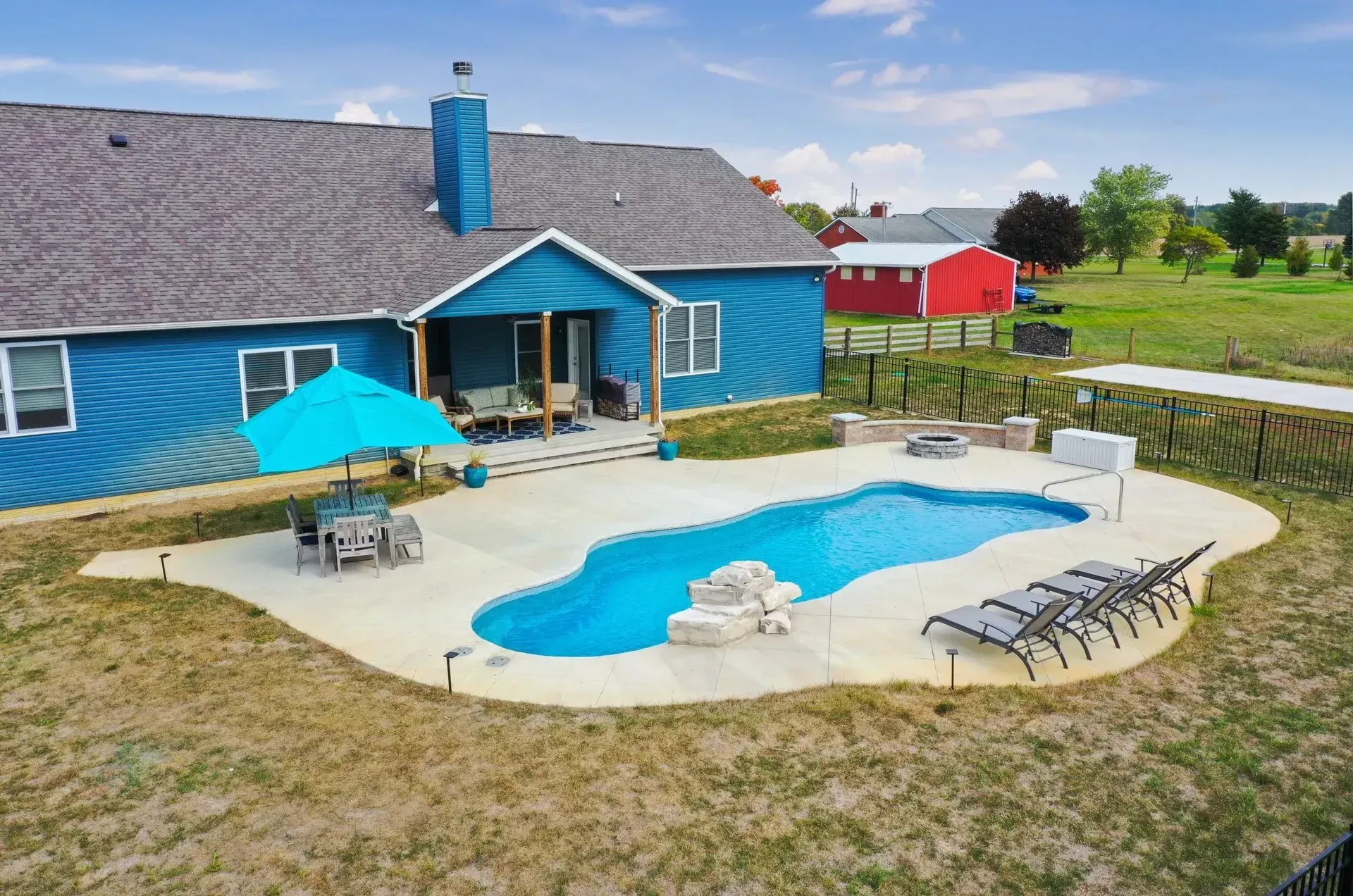 Blue house with a backyard pool, umbrella, and lounge chairs on a sunny day. Fields and a red barn are in the background.