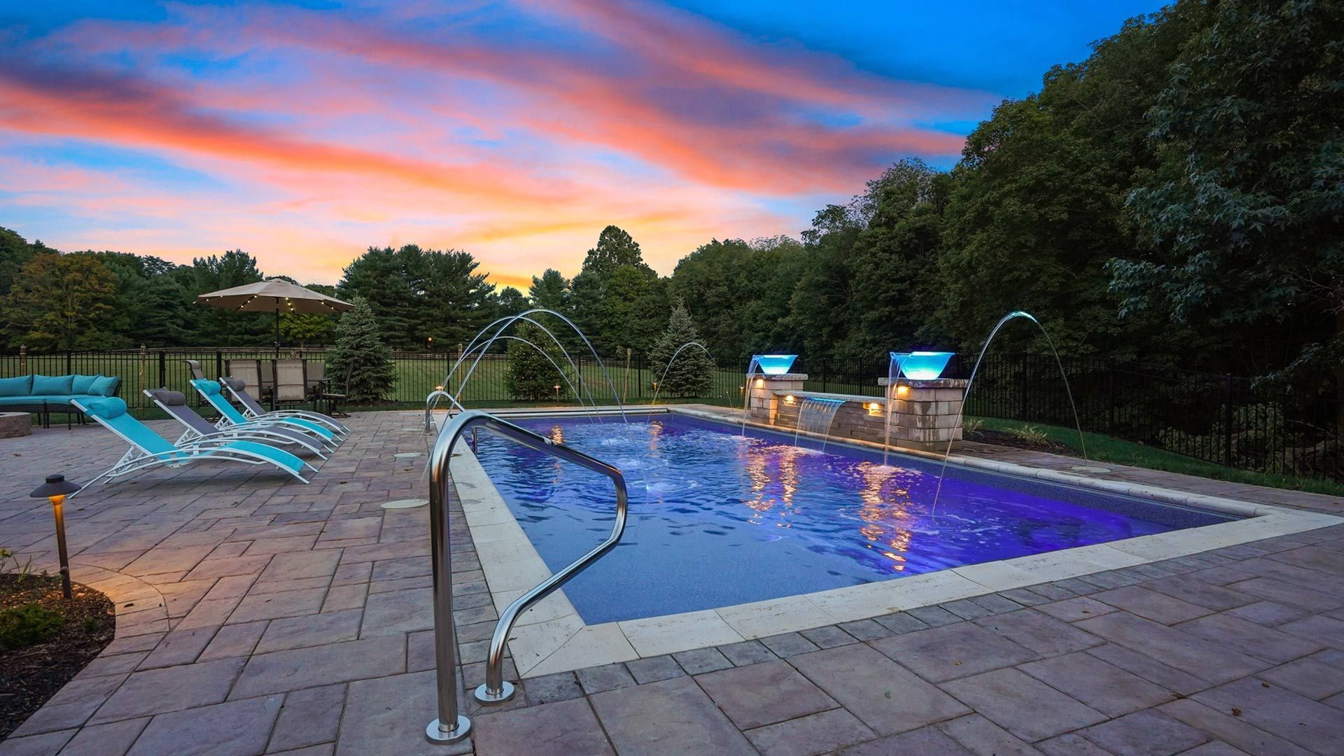 Sunset view of a rectangular pool with blue lights and water fountains. Turquoise lounge chairs sit on a stone patio.