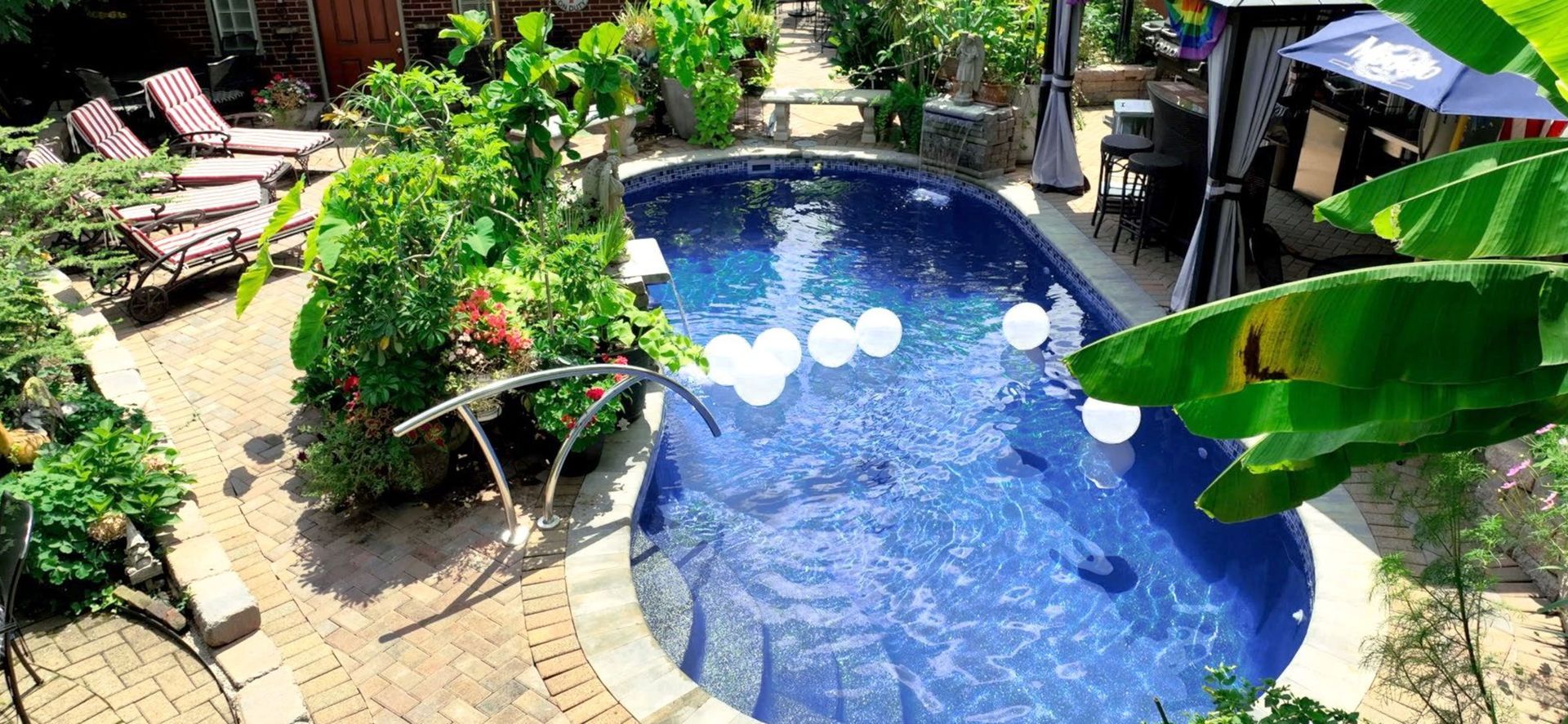 A backyard pool with white balloons floating in the water, surrounded by lush greenery and brick paving. Lounging chairs sit to the left.