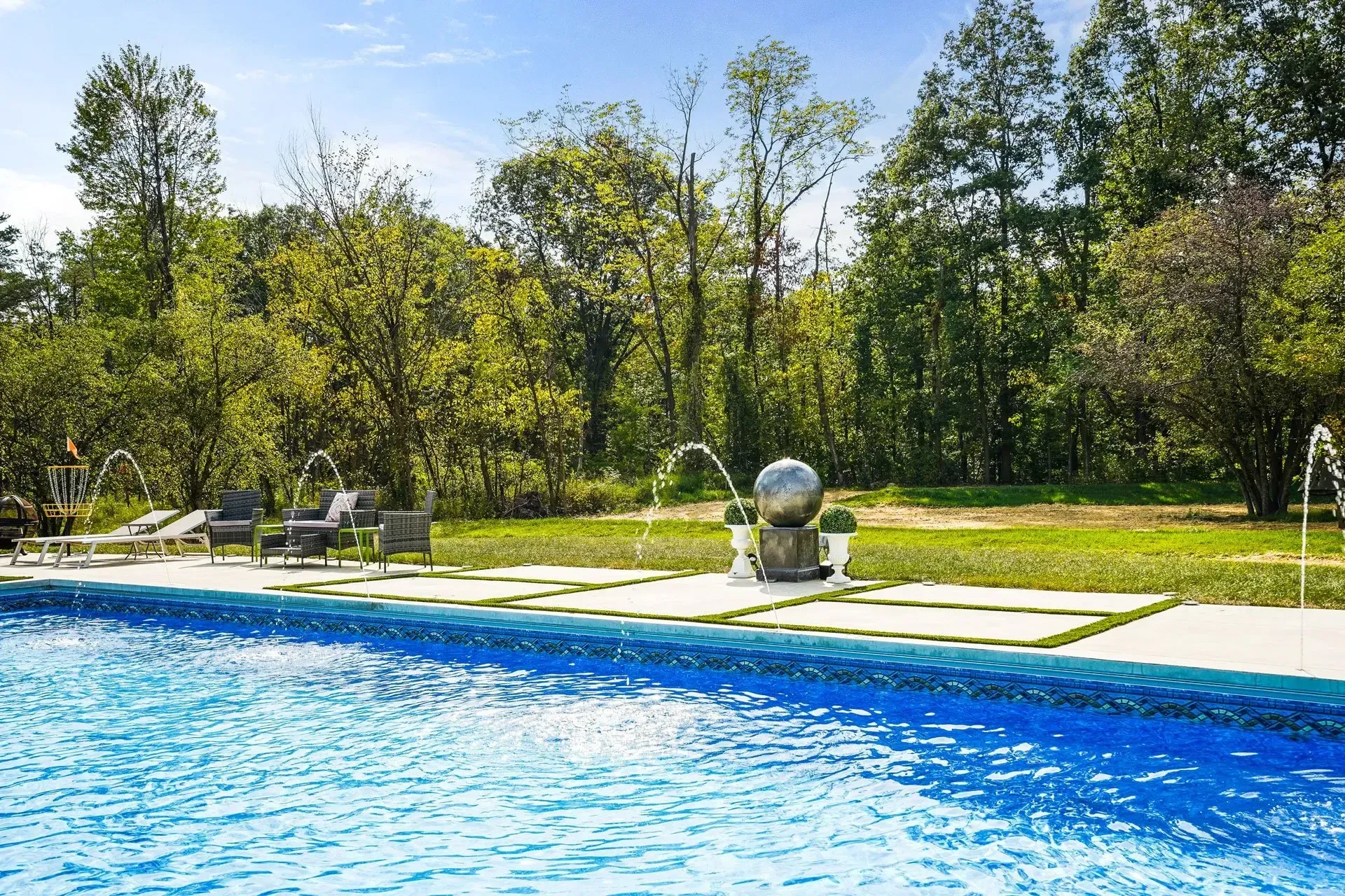 A luxurious outdoor pool with fountains and a modern sphere sculpture, surrounded by a green lawn and trees on a sunny day.