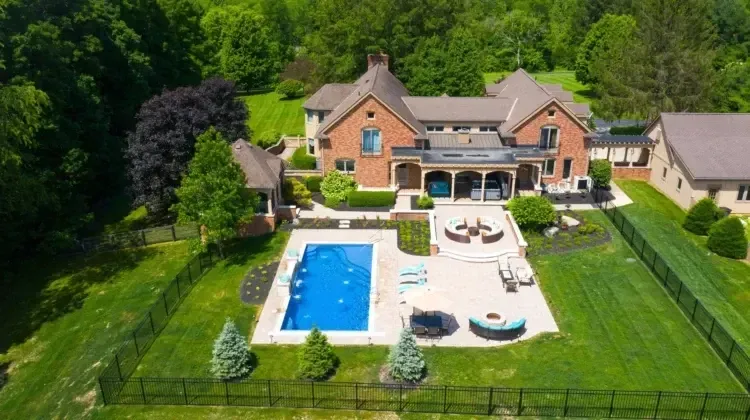 Aerial view of a luxurious brick mansion with a pool and patio area surrounded by green grass and trees.