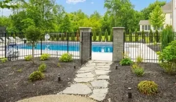 Stone path leads to a pool area with black fence, flanked by landscaping and green trees, on a sunny day.