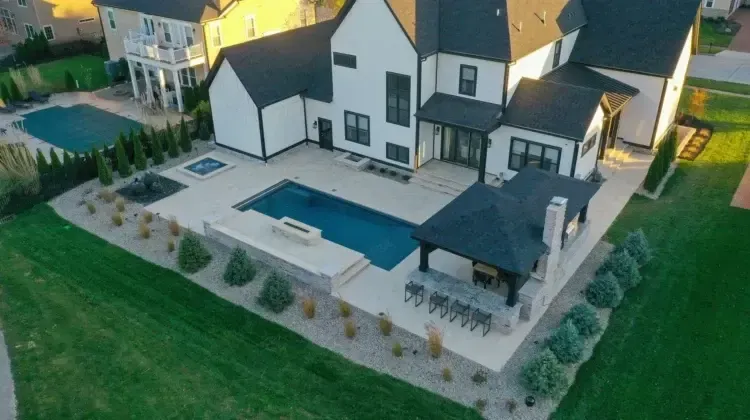 Aerial view of a modern white house with black trim, a pool, patio, and a gazebo on green grass.