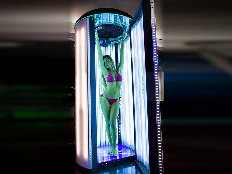 Donna in bikini rosa in piedi in una cabina abbronzante verticale. Luci fluorescenti la illuminano.