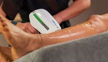 Persona che utilizza un dispositivo portatile su una gamba, probabilmente per la depilazione. Il dispositivo è bianco con dettagli verdi e la pelle appare lucida.