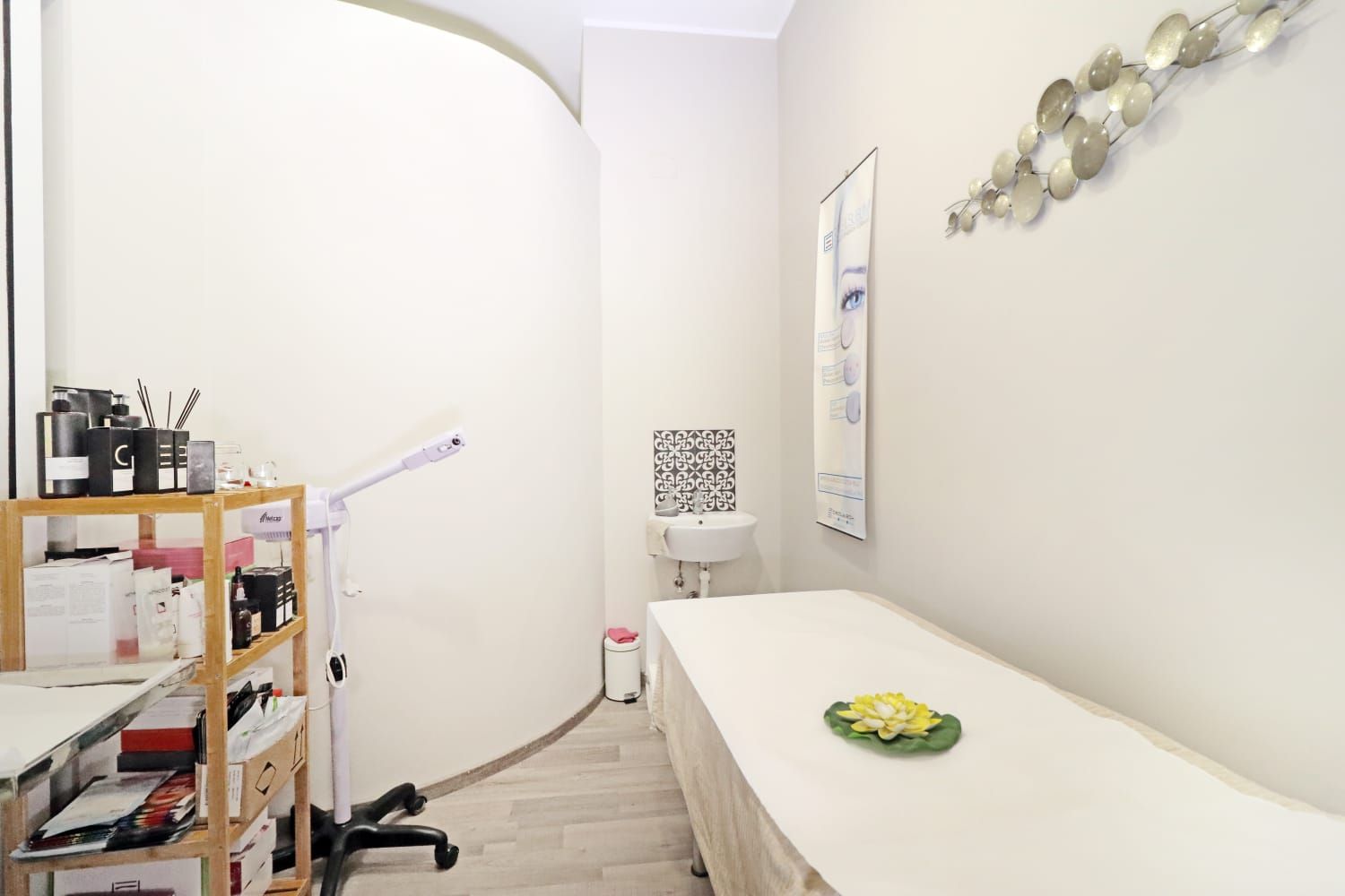 Sala trattamenti spa con letto, lavandino, scaffali con prodotti e quadri decorativi alle pareti.