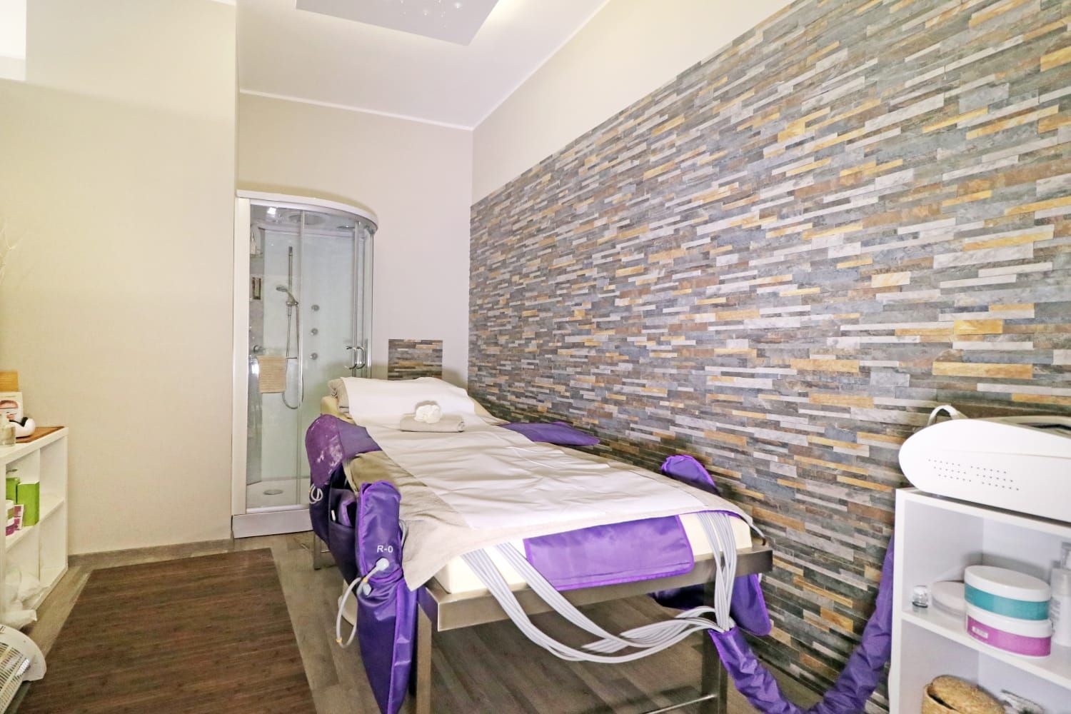 Sala spa con lettino per massaggi, parete con elementi in pietra, doccia a vista. Dettagli viola, biancheria bianca e mensole.