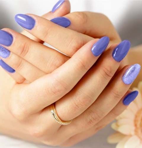 Mani con unghie lunghe e ovali viola, una delle quali è glitterata. Anello d'oro a un dito. Sfondo chiaro.