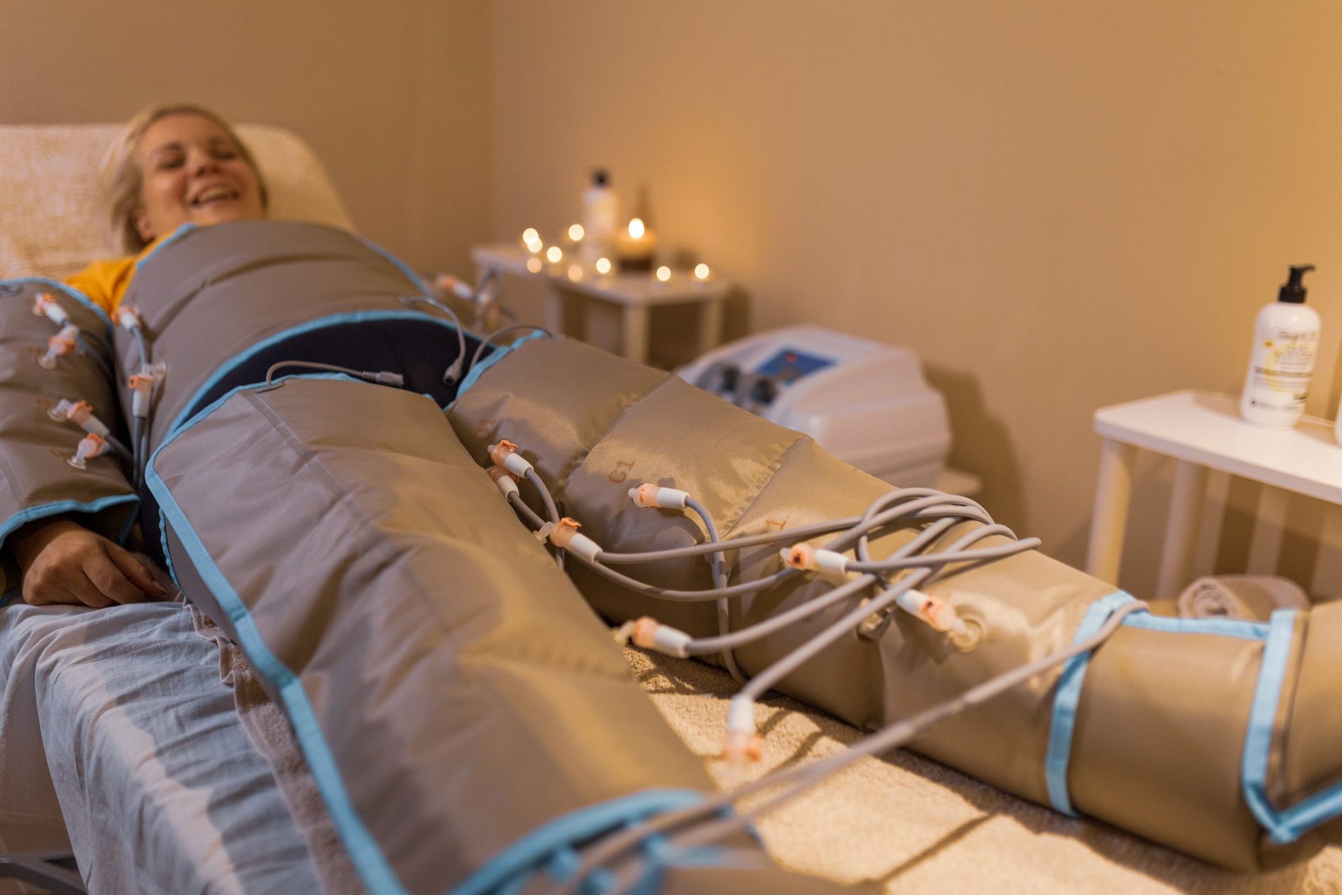Donna sdraiata su un letto con maniche a compressione, sorridente. Ambiente spa con candele e attrezzature.