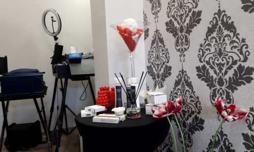 Una postazione trucco con un tavolo nero con prodotti di bellezza, fiori e un centrotavola con bicchiere da martini, accanto a una parete con un motivo damascato.