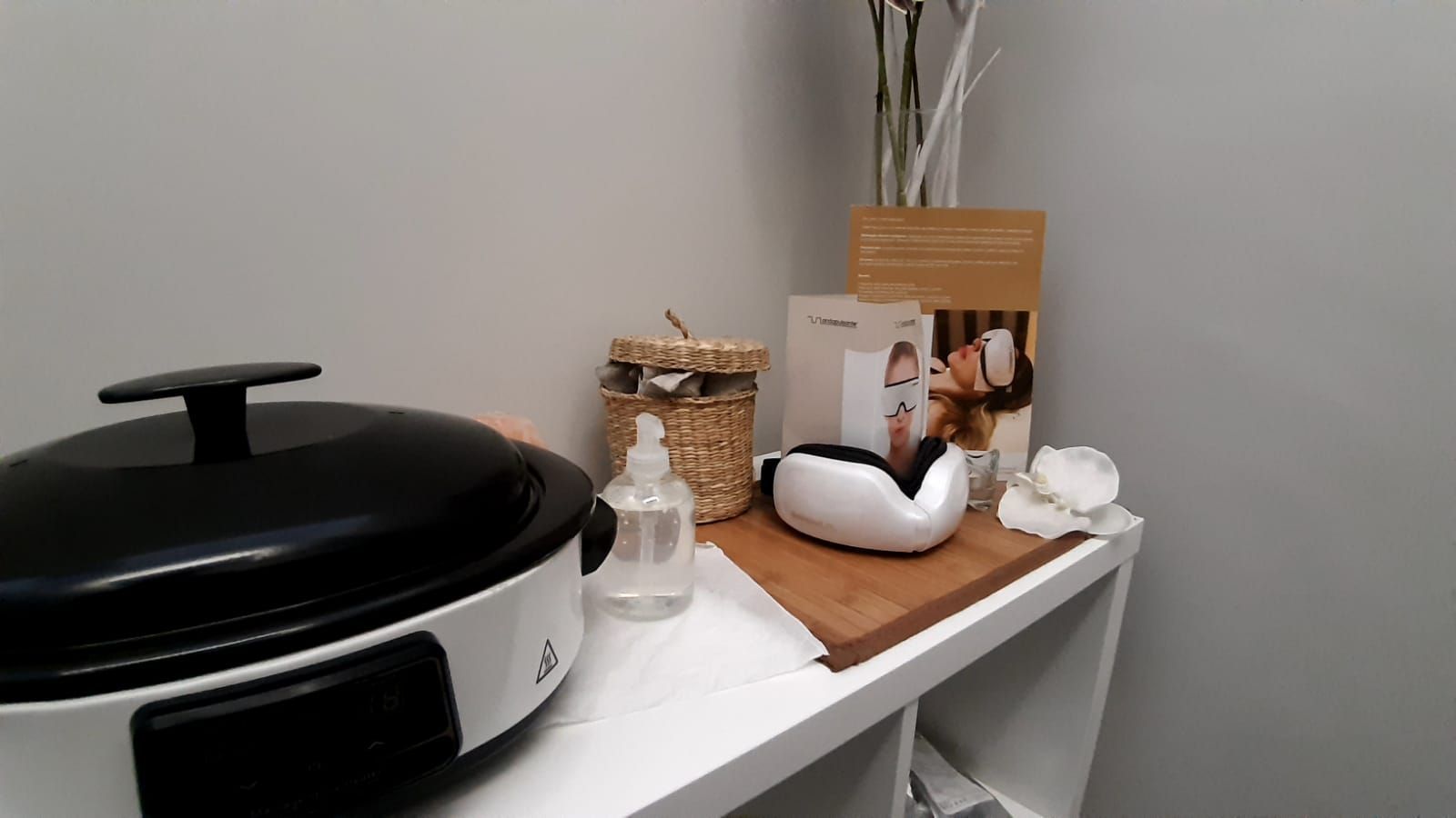 Un allestimento da spa con un dispositivo di riscaldamento con coperchio nero, un dispositivo per massaggi e decorazioni su uno scaffale bianco.