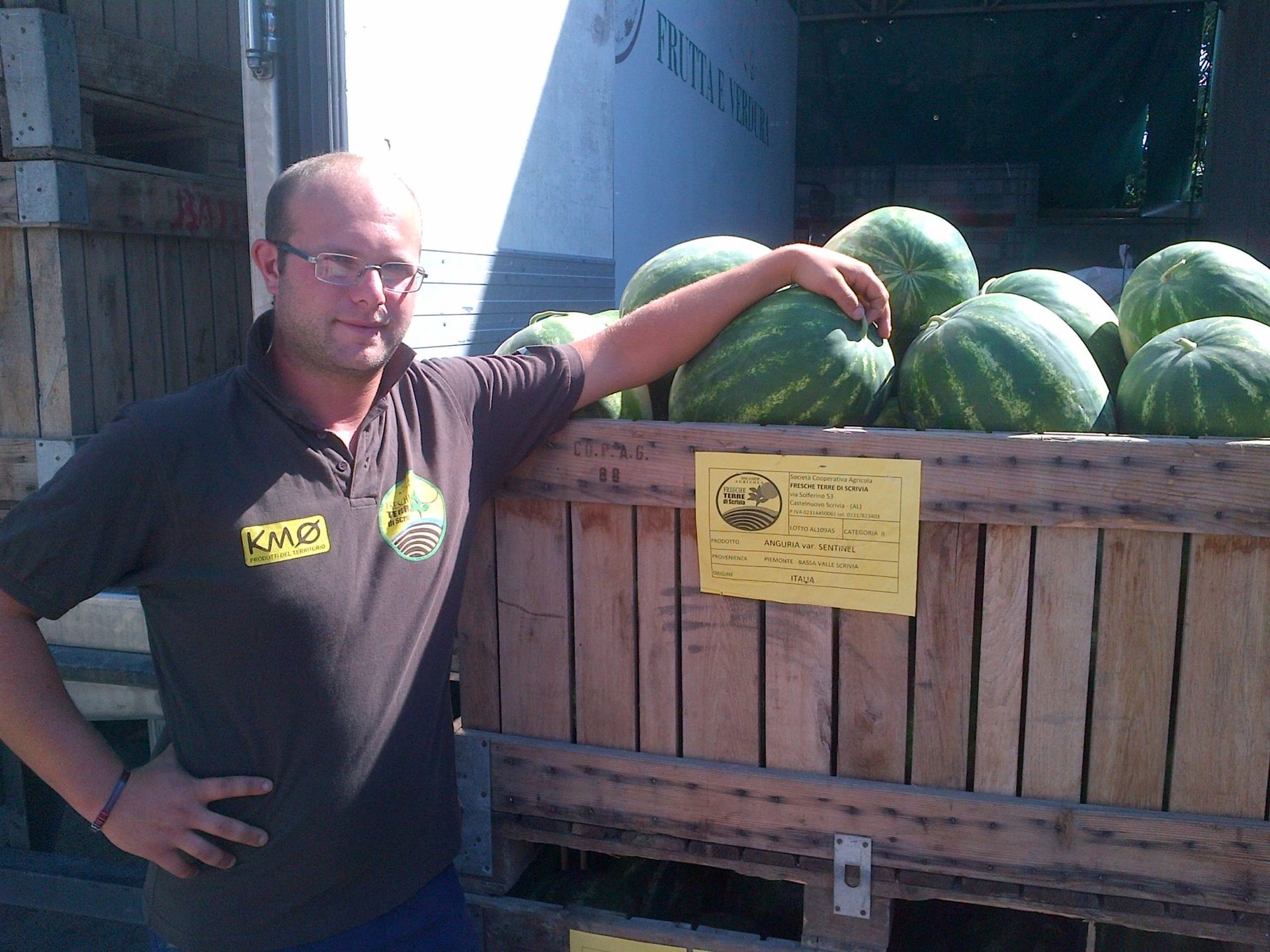Un uomo con una camicia marrone è appoggiato a una cassa di legno piena di angurie.