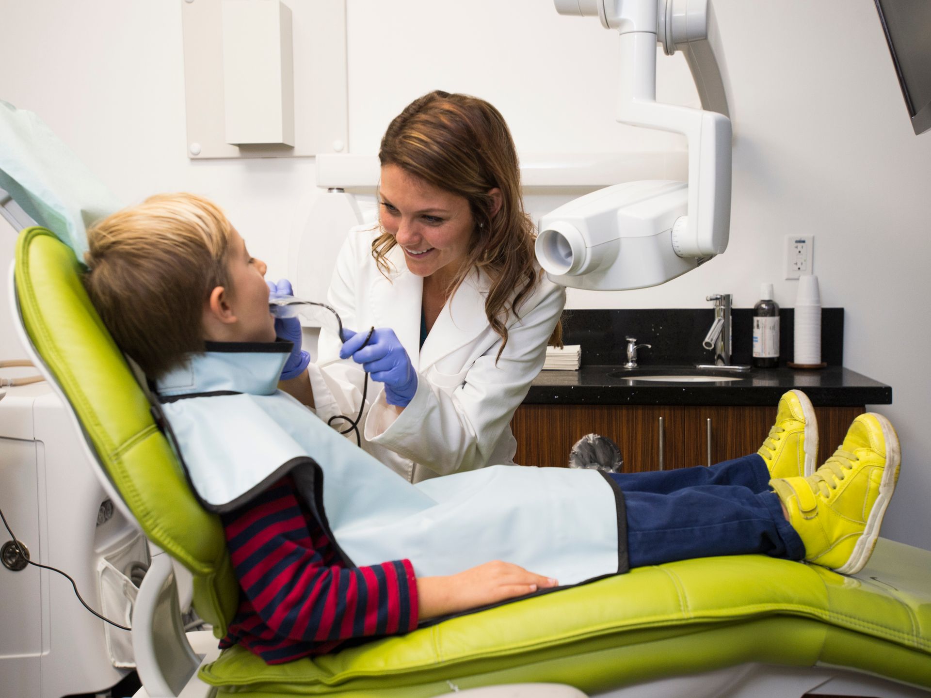 A young boy is sitting in a pediatric dentist's chair while a male dentist is checking his teeth.