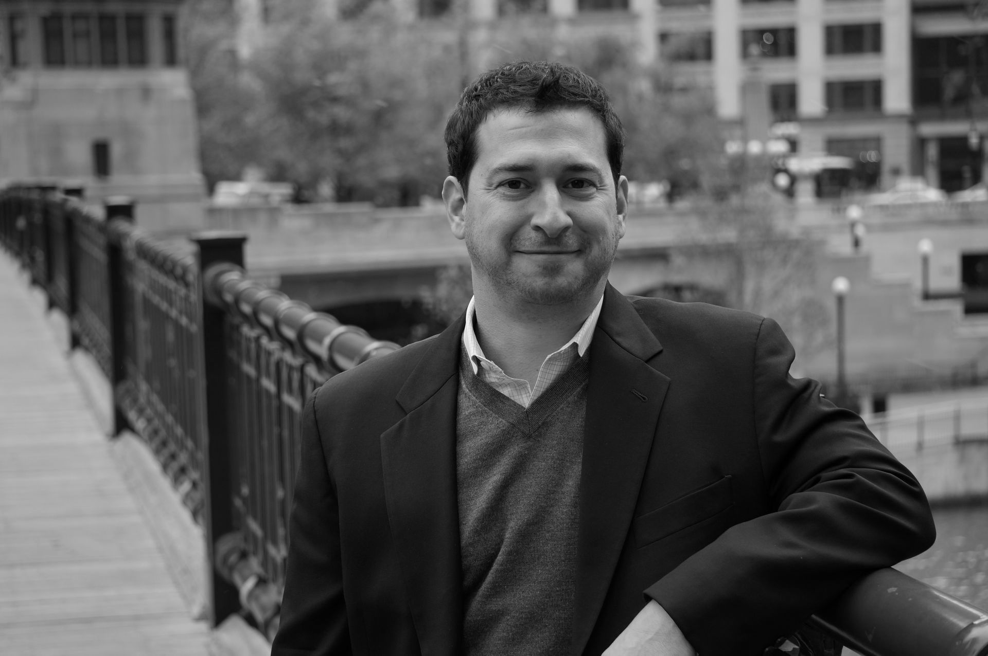 A man in a suit and sweater is leaning on a railing on a bridge.