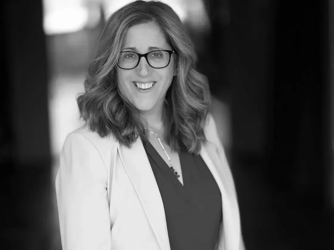 A woman wearing glasses and a white jacket is smiling in a black and white photo.