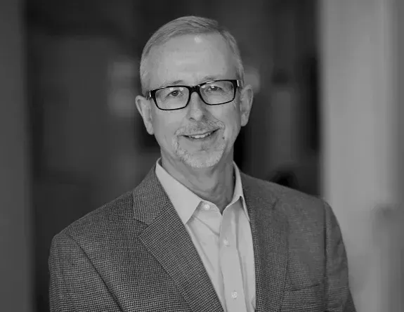 A man in a suit and glasses is smiling in a black and white photo.