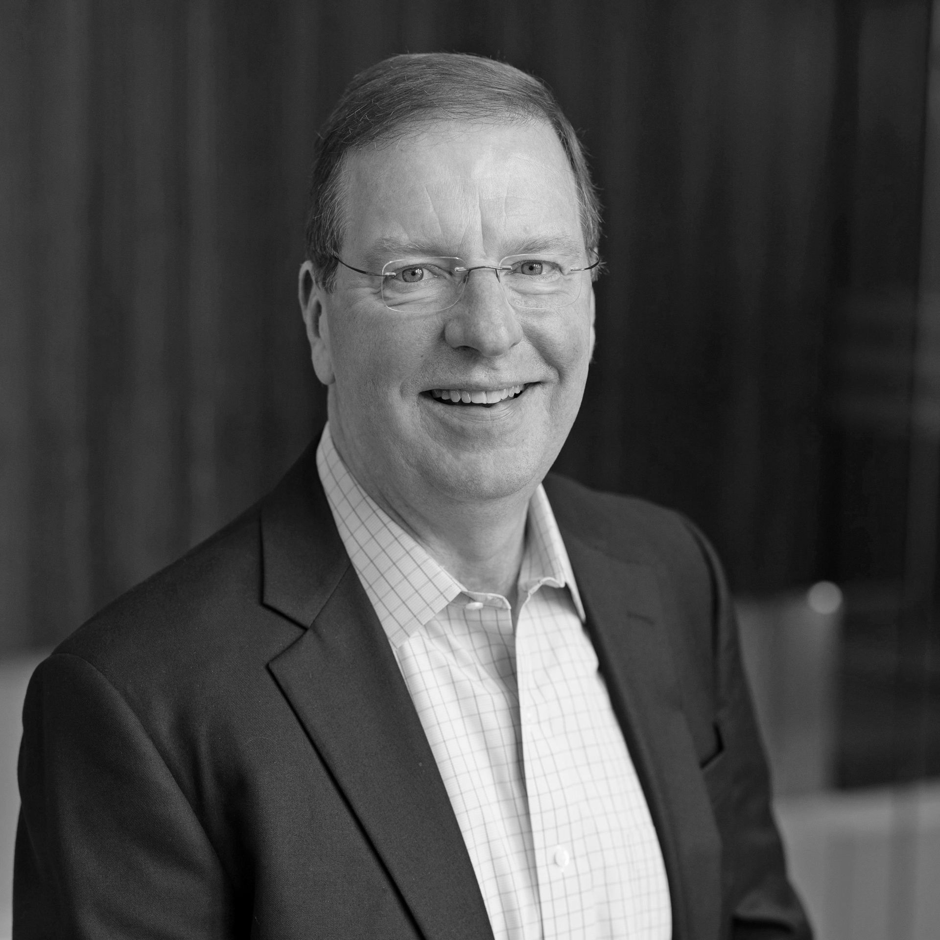 A man in a suit and glasses is smiling in a black and white photo.