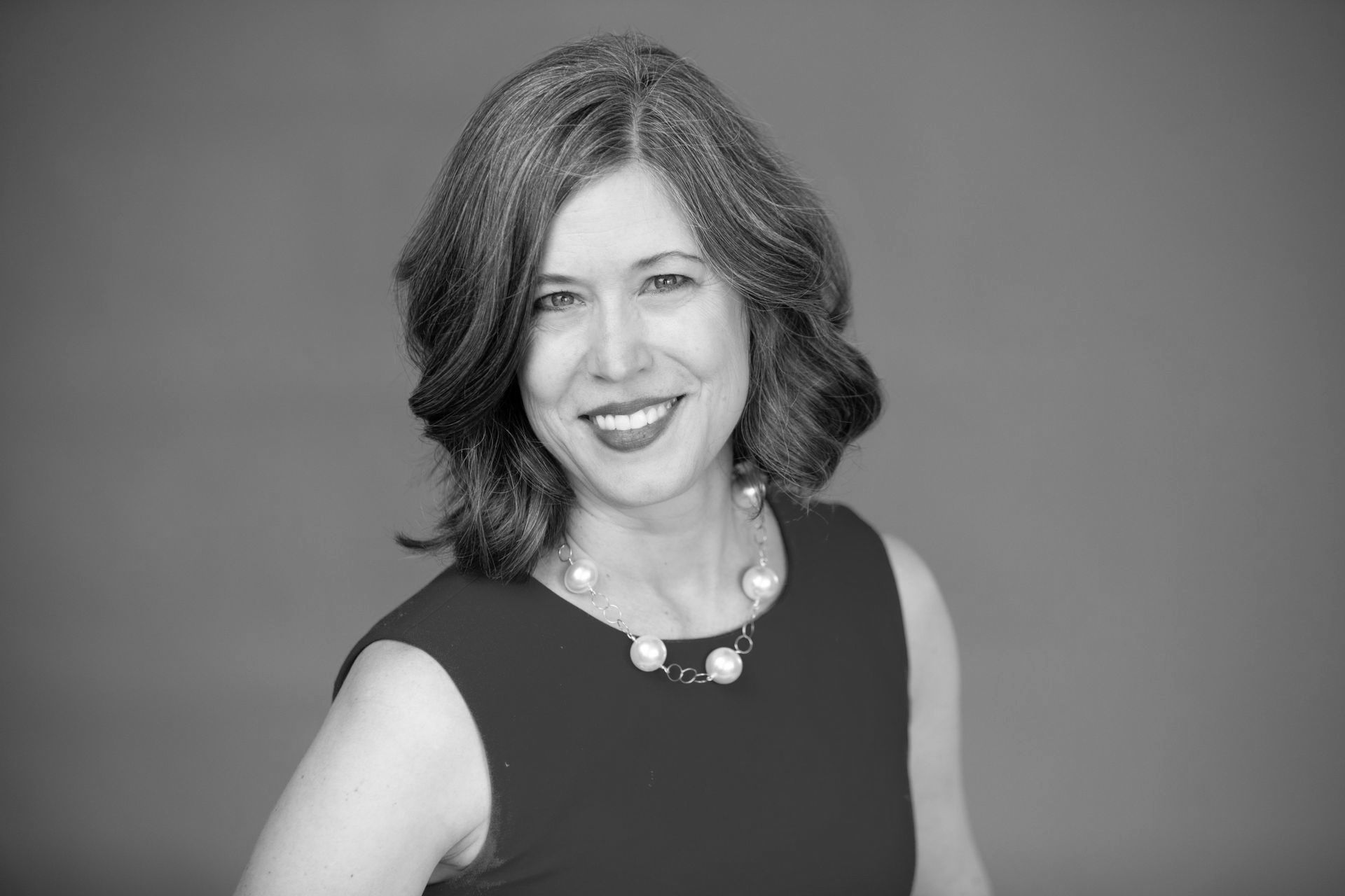 A woman in a black dress and pearl necklace is smiling in a black and white photo.