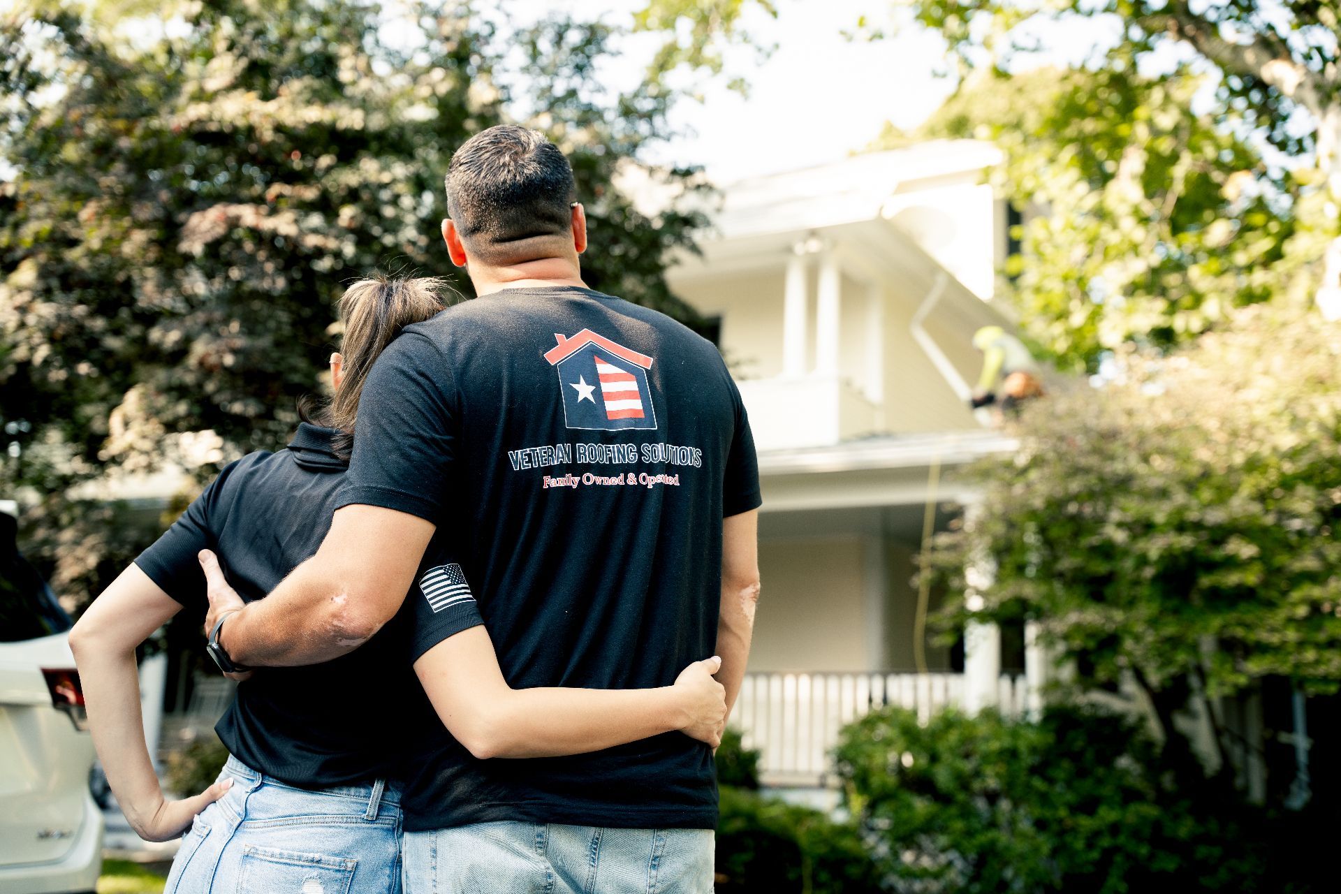 Couple embraces, gazing at a white house with a porch and green foliage. Man wears a shirt with a logo.